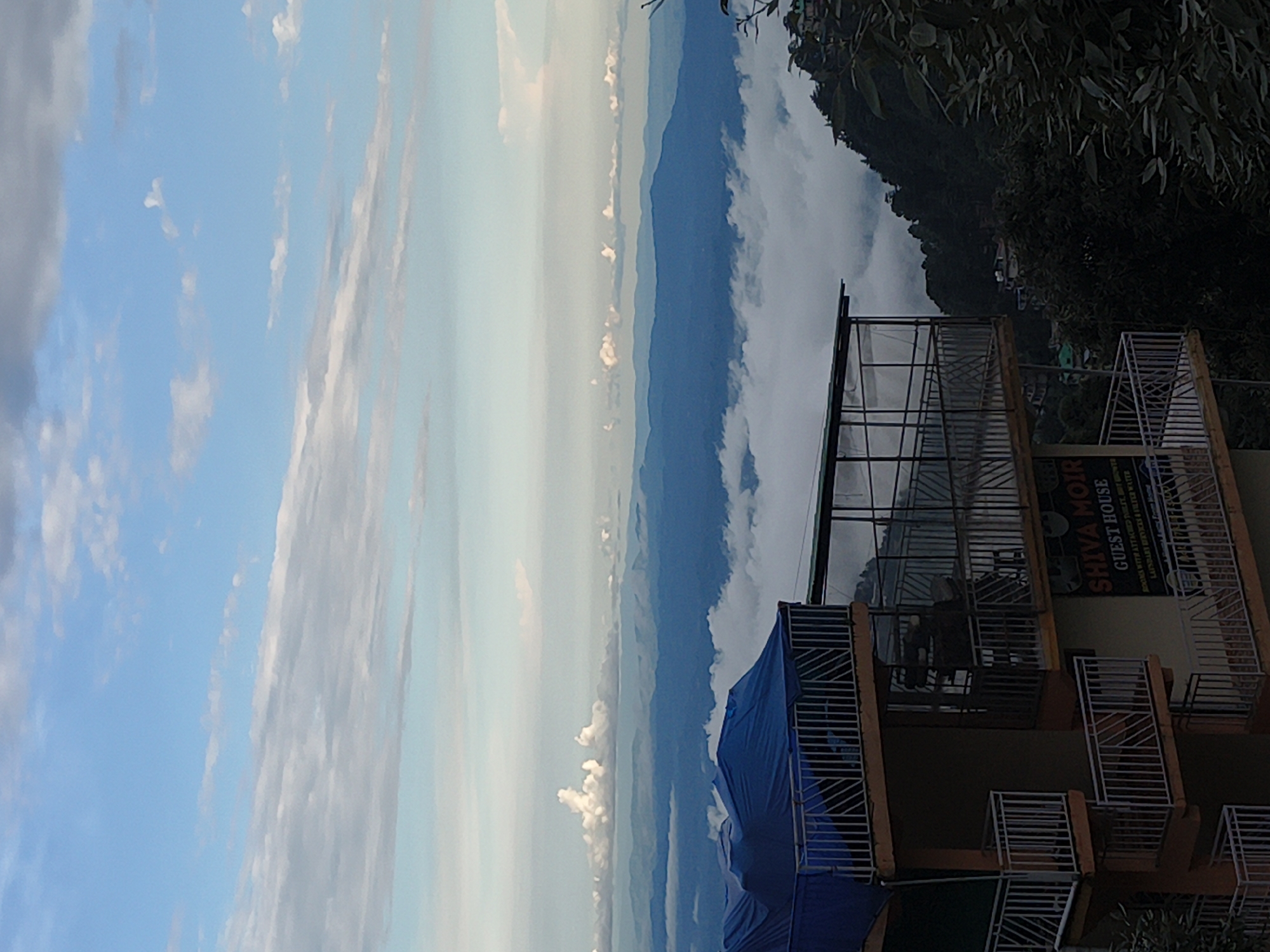 The image shows a multi-story guesthouse building with balconies, situated on a hillside overlooking a valley filled with clouds. In the foreground, the building's balconies are visible, some covered with a blue tarp. A sign on the building reads "SHIVA MOIR GUEST HOUSE". Beyond the building, layers of mountains recede into the distance, shrouded in a soft, atmospheric haze. The sky above is a mix of blue with scattered white clouds. The environment appears to be a mountainous region, possibly in India, given the reference to Haini, India in the prompt. There are no people visible in the image.