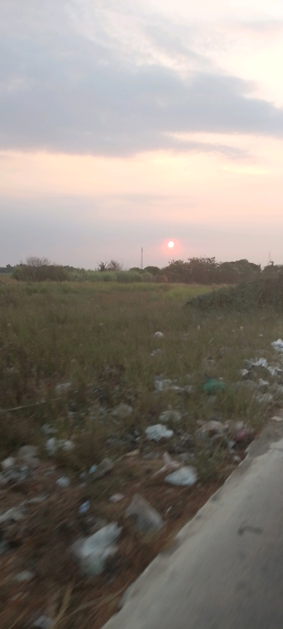 The image depicts a landscape scene, likely at dusk or dawn, with a muted color palette. The sky is a blend of soft grays and pinks, with a faint, circular sun visible in the upper right quadrant. Below the sky is a low horizon line, dotted with sparse trees and vegetation. The foreground is characterized by a field of dry grass and scattered debris, including what appears to be plastic bags and other refuse. A concrete structure, possibly a road or sidewalk edge, is visible along the right side of the frame. The overall impression is one of a rural or semi-rural area, possibly experiencing some environmental neglect due to the presence of litter. This scene is located in Sukaraja, Indonesia.