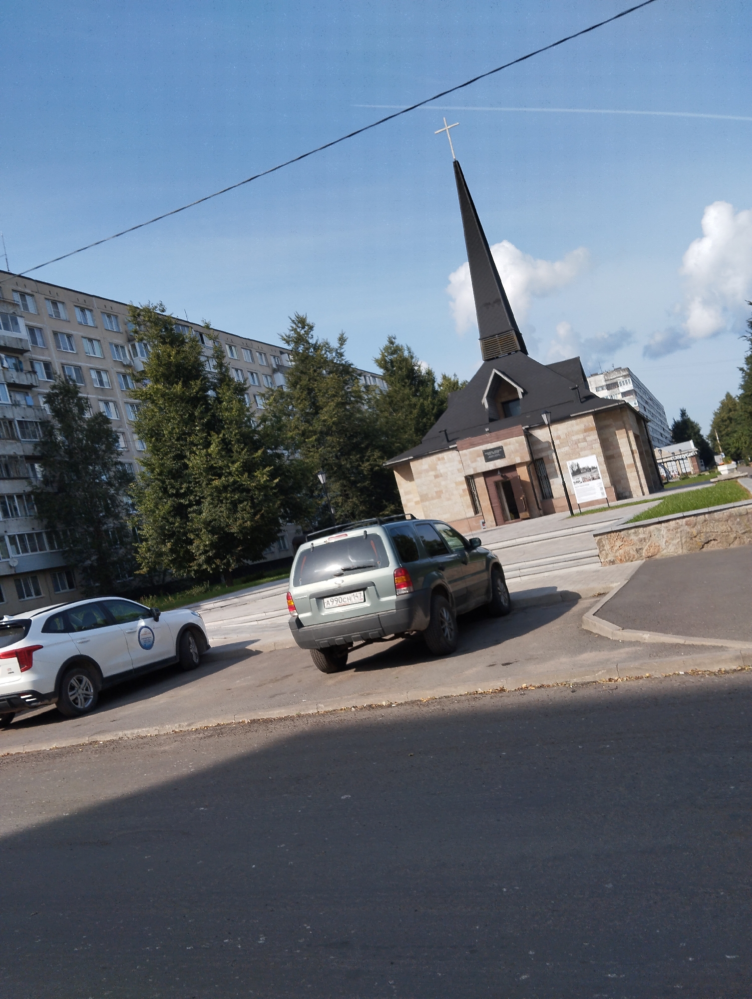 The image depicts a street scene in Kingisepp, Russia, under a partly cloudy sky. In the foreground, a dark asphalt road is visible with a distinct shadow falling across it. To the left, a white SUV with a blue circular decal on its side is parked, partially obscuring a light-colored building with many windows. To the right of the white SUV, a dark-colored SUV is also parked.
Dominating the mid-ground is a modern church structure with a distinctive dark, pointed spire topped with a white cross. The church building itself is constructed of light-colored stone. In the background, a large, multi-story apartment building with numerous windows stretches across the frame. Lush green trees are interspersed between the apartment building and the church. A dark power line stretches diagonally across the upper portion of the image. The overall impression is a blend of residential, religious, and public spaces within a Russian urban environment.