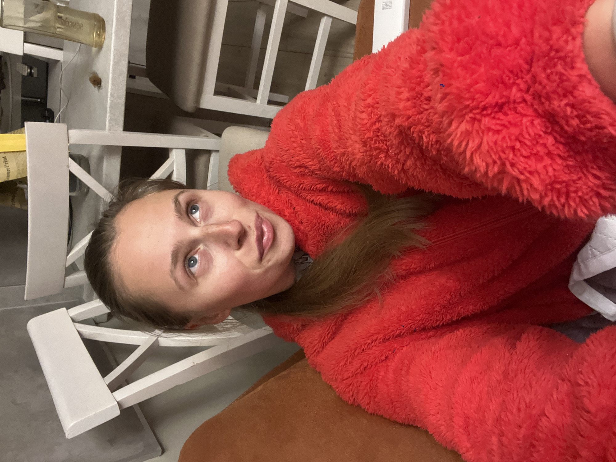 A woman with blonde hair is wearing a red fuzzy jacket while sitting on a brown chair, possibly indoors. In the background, there is a white chair and a bottle on a table. The location is not identifiable as Rafailovići, Montenegro from the information given.