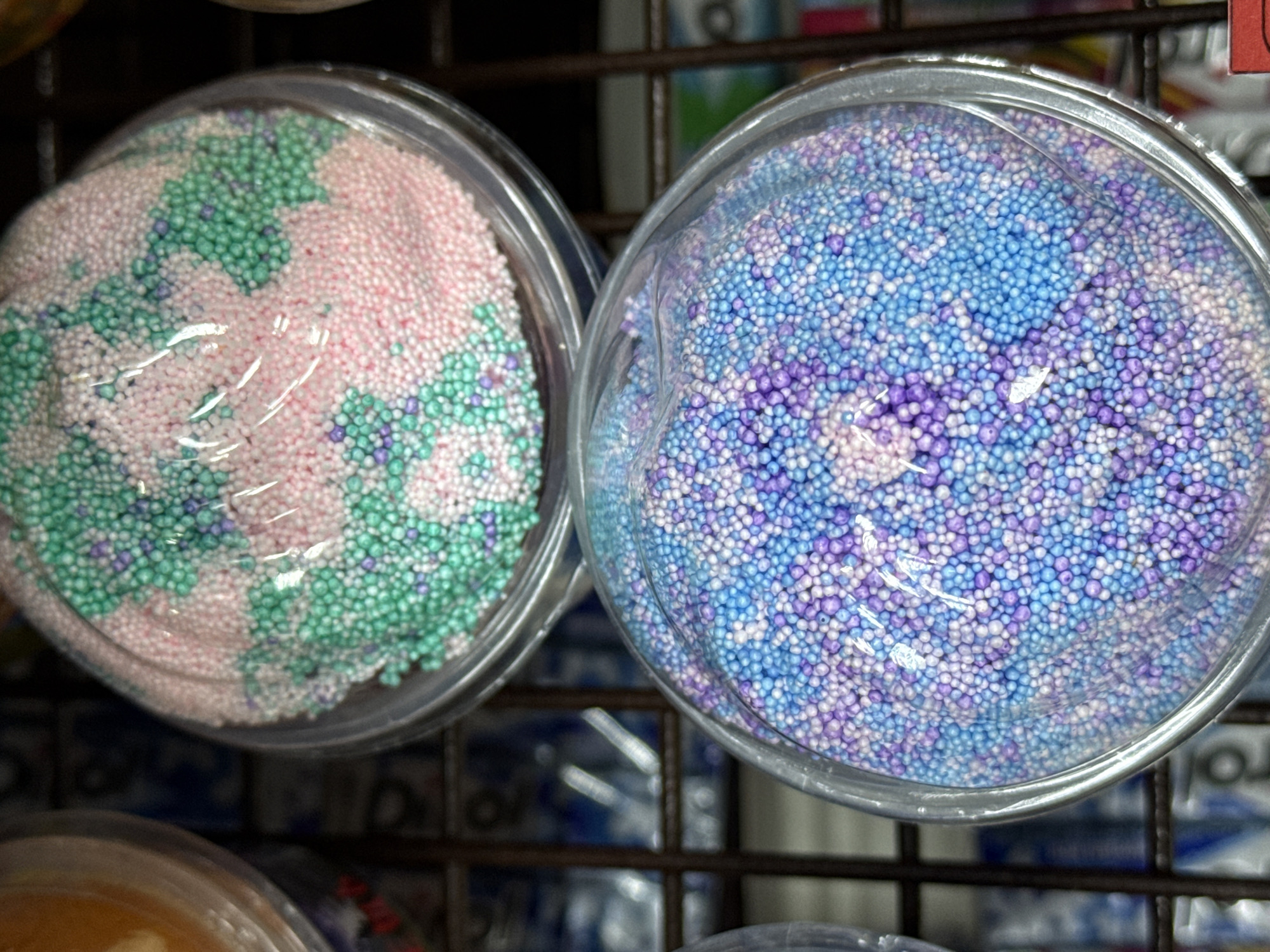 Two clear plastic containers are filled with small, colorful foam balls. The container on the left holds pink, green, and white balls. The container on the right holds blue, purple, and white balls. Both containers appear to be displayed on a metal shelf. The exact location within Votkinsk, Russia, is not discernible.