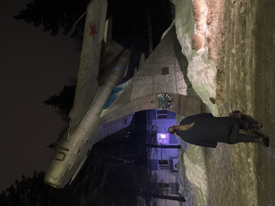 A nighttime exterior photograph taken in Klin, Russia, depicts a female individual walking towards a large aircraft monument during winter. The primary subject is a light-colored, Soviet-era fighter jet, identified as a MiG-15, marked with the number "01" on its fuselage and a red star insignia on its right wing. The aircraft is angled upwards on a textured silver pedestal featuring a blue section and a plaque with Cyrillic text that translates to "Dedicated to the pilots of our Air Defense." Small floral tributes are visible at the base of the pedestal. The ground is covered with snow, forming uneven piles, with a partially cleared pathway in the foreground. In the left background, a low-rise, light-colored building with multiple windows is visible; one first-floor window emits a prominent pink-purple light. Dark evergreen trees rise against the dark, overcast sky in the background. The individual, viewed from behind, has long light-brown hair in a ponytail and wears a long black coat, dark pants, and dark boots. The scene is illuminated by artificial light sources.