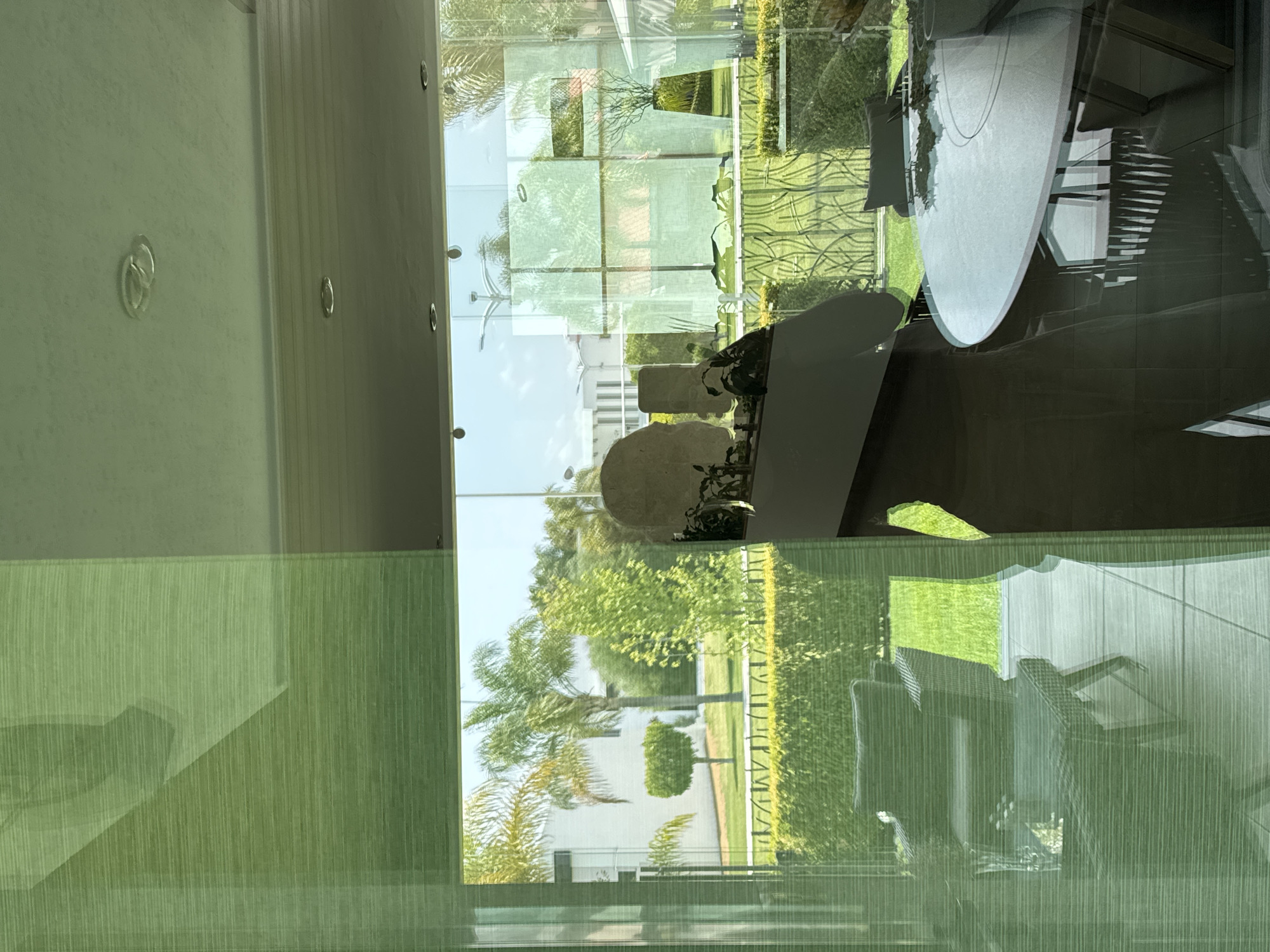 The image displays an interior view looking out through a large window. The window glass has a faint green tint and fine vertical lines, possibly from integrated blinds or textured glass. The outdoor scene is a brightly lit, landscaped area under a clear, blue sky.
Outside the window, lush green grass stretches across the ground, interspersed with neatly trimmed hedges and a variety of trees, including prominent palm trees. A white building is visible in the distance, partially obscured by the foliage. A decorative metal fence or railing with an ornate, wavy design can be seen.
Reflected on the window's surface are elements from the interior space. These reflections include a large, round, light-colored table, parts of what appear to be chairs or furniture, and a staircase with visible steps. Also reflected are a light-colored, possibly stone-like object, and some potted plants. Inside, on a white wall adjacent to the window, multiple circular, recessed ceiling lights and decorative ring-shaped fixtures are visible. A section of a white ceiling with linear paneling is also present.
The image depicts a sunny daytime with clear weather. No people are present, and no specific activity is taking place. There is no visible text in the image. The setting suggests a modern architectural space with a well-maintained outdoor environment.