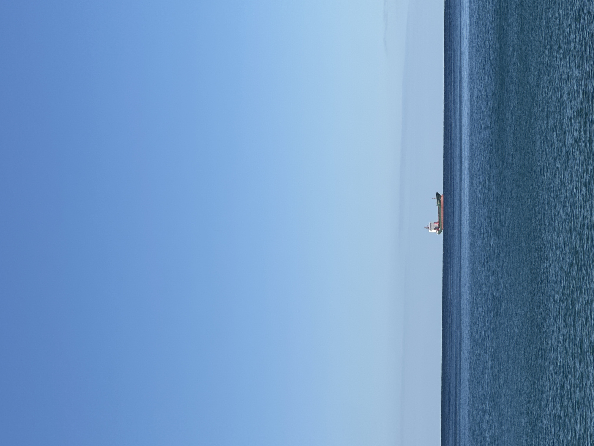 The image shows a horizontal view of the sea and sky in Šušanj, Montenegro. The sky is a gradient of blue, lighter near the horizon. The sea is a darker shade of blue with visible water ripples. A cargo ship is seen on the horizon, positioned at the vertical center of the frame.