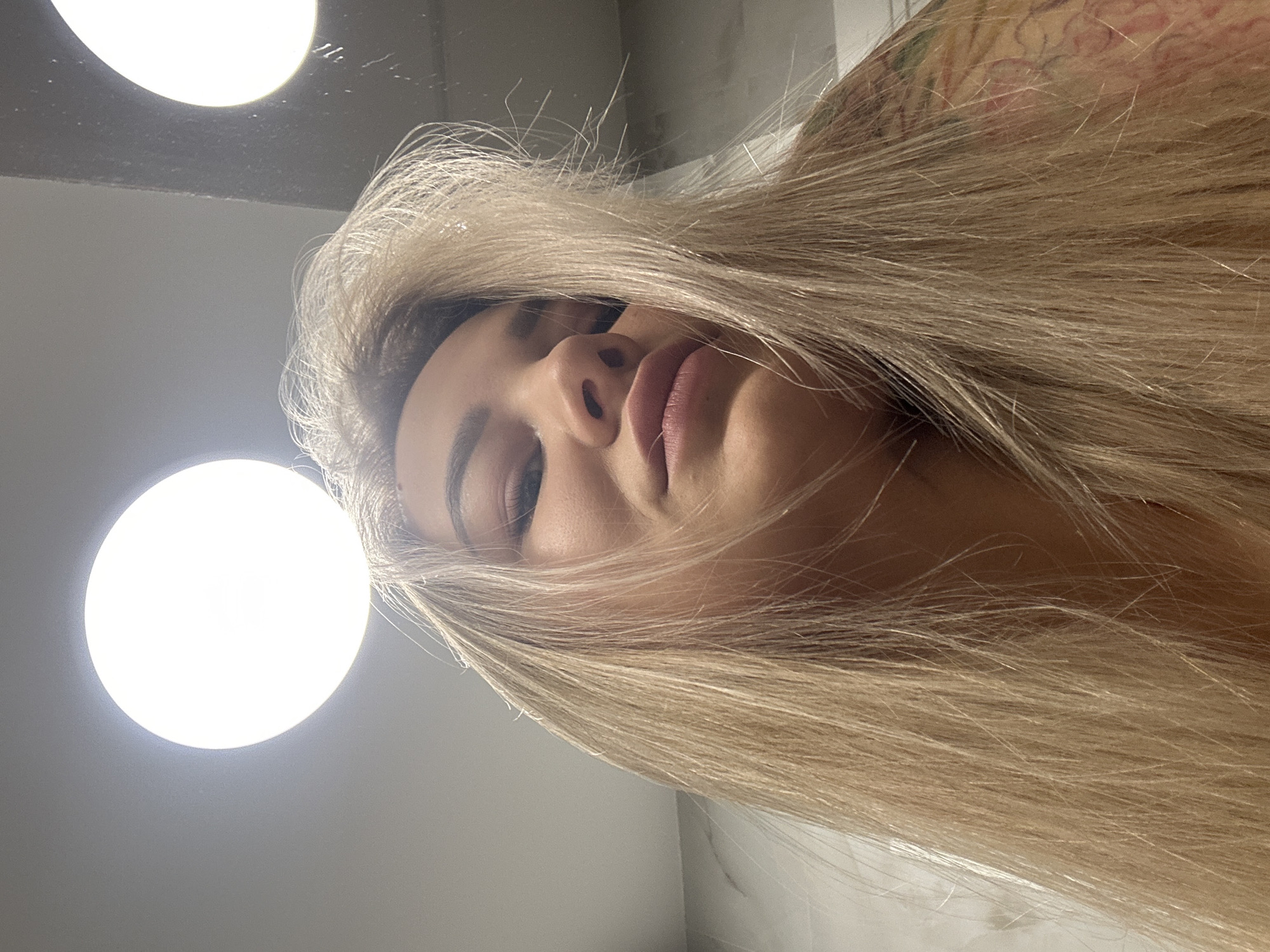 The image is a low-angle shot of a woman with long, light blonde hair. The woman's face is prominently featured, with her eyes partially closed and a slight tilt of her head. The lighting is bright, with two circular light fixtures visible in the background. The background appears to be a room with light-colored walls. There is no information available to determine if this image was taken in Budva, Montenegro.