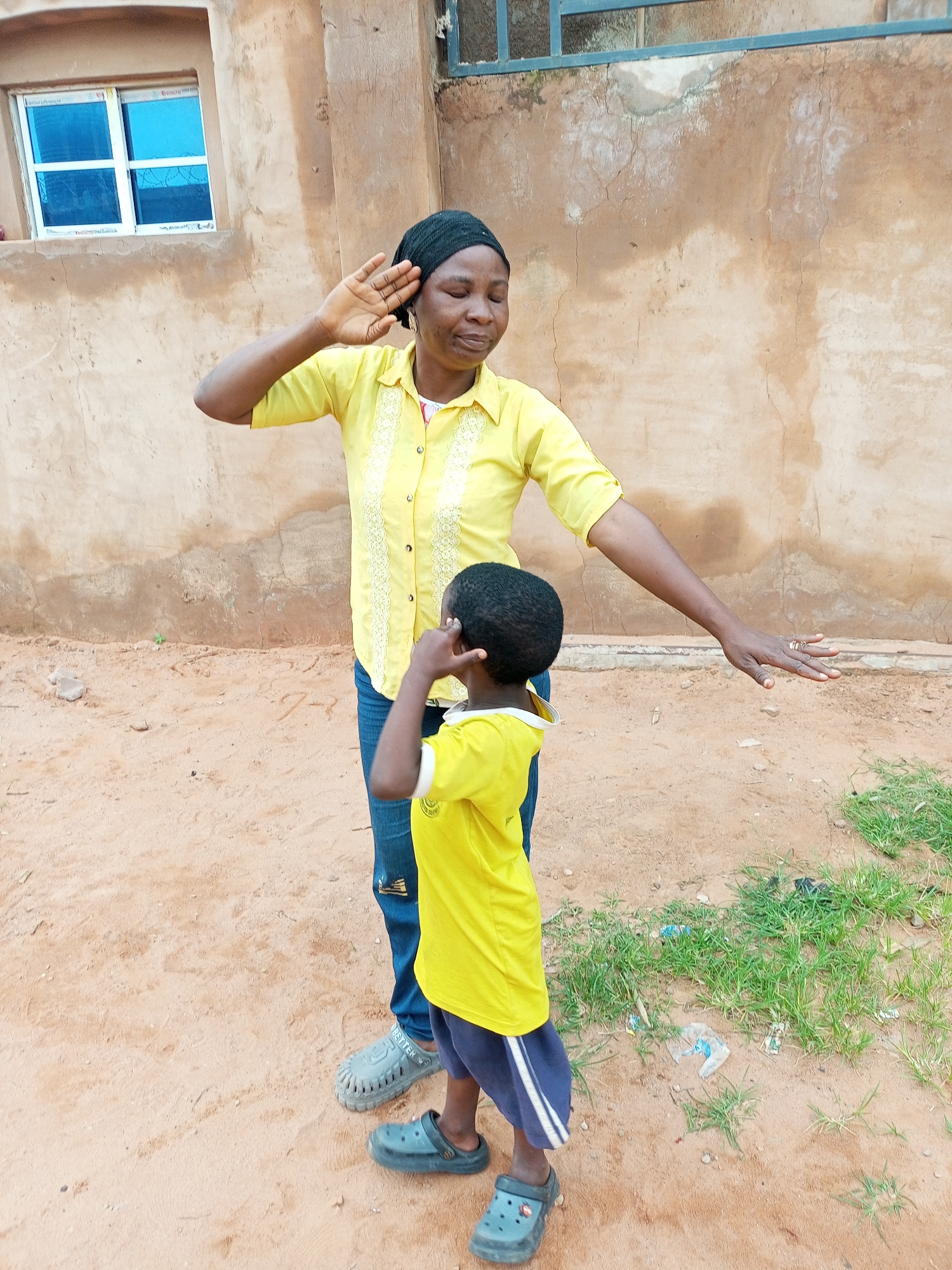 The image depicts two individuals, an adult woman and a child, standing on a dirt ground in front of a textured, weathered wall. The woman, wearing a yellow shirt and a black head covering, has her right arm raised with her hand near her forehead, and her left arm extended to her side. She appears to be looking downwards. The child, facing away from the camera, is wearing a yellow shirt and dark shorts with white stripes. The child's right hand is raised to their face.
The environment suggests an outdoor setting, possibly a courtyard or an open area adjacent to a building. In the background, there's a building with a brown, uneven wall and a window with blue panes. Patches of green grass are visible on the right side of the frame, with some scattered debris. The ground is primarily composed of sandy soil with visible footprints and small stones. The overall impression is a casual, everyday scene in a rural or semi-urban environment. The location is identified as Tempure, Nigeria.