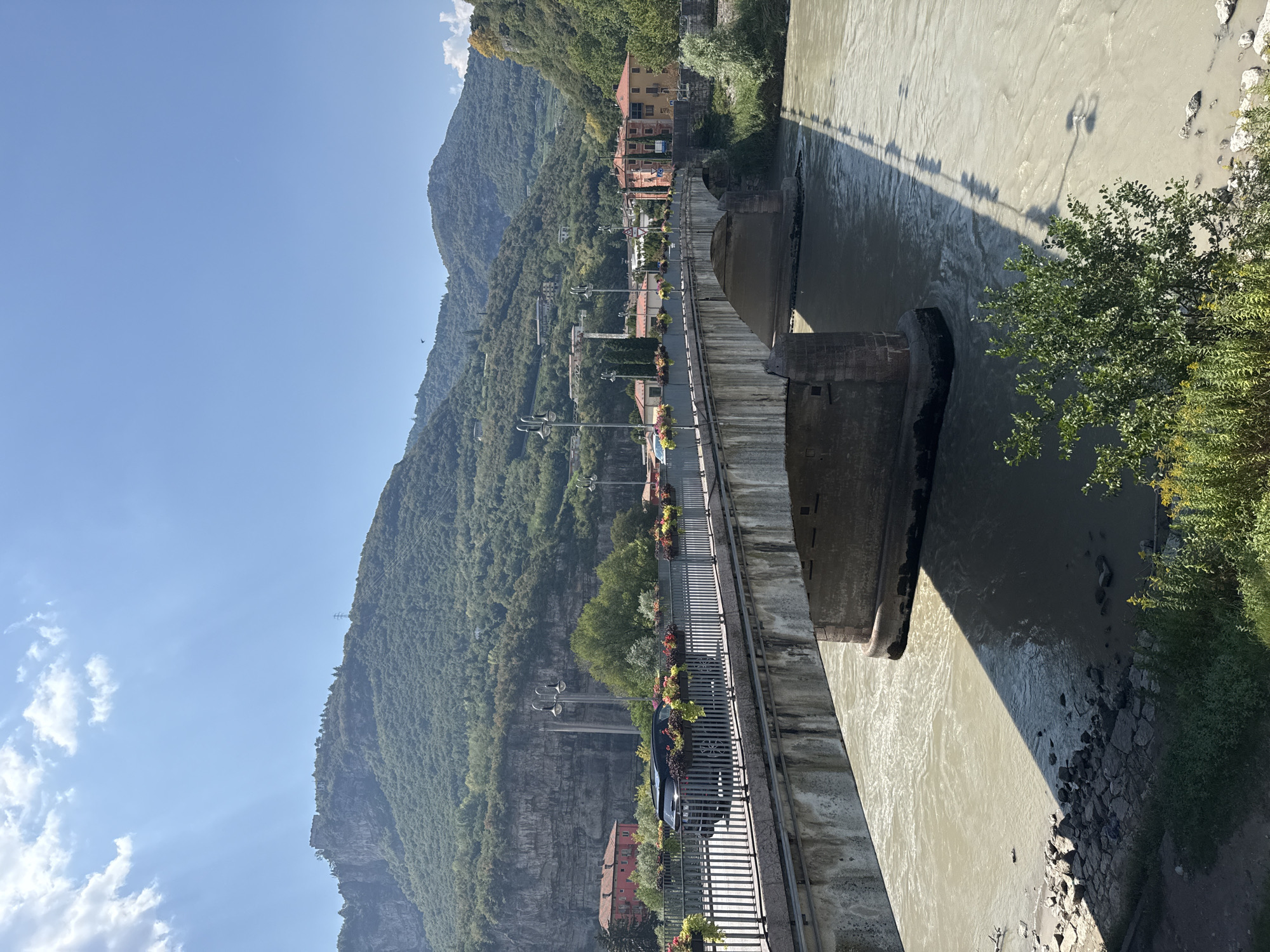 The image depicts a wide, murky river flowing under a concrete and stone bridge. The bridge is lined with decorative planters filled with colorful flowers and has a metal railing. A dark-colored SUV is driving across the bridge. On the far bank of the river, there are buildings with orange and yellow facades, surrounded by trees. In the background, steep, tree-covered mountains rise, with some rocky outcrops and what appears to be ruins or a small structure at the summit of one of the peaks. The sky is a clear blue with a few scattered white clouds, and the sun is casting a distinct shadow of the bridge onto the water. The overall scene suggests a sunny day in a town nestled within a mountainous landscape, likely Trento, Italy, given the architectural style and mountainous terrain.