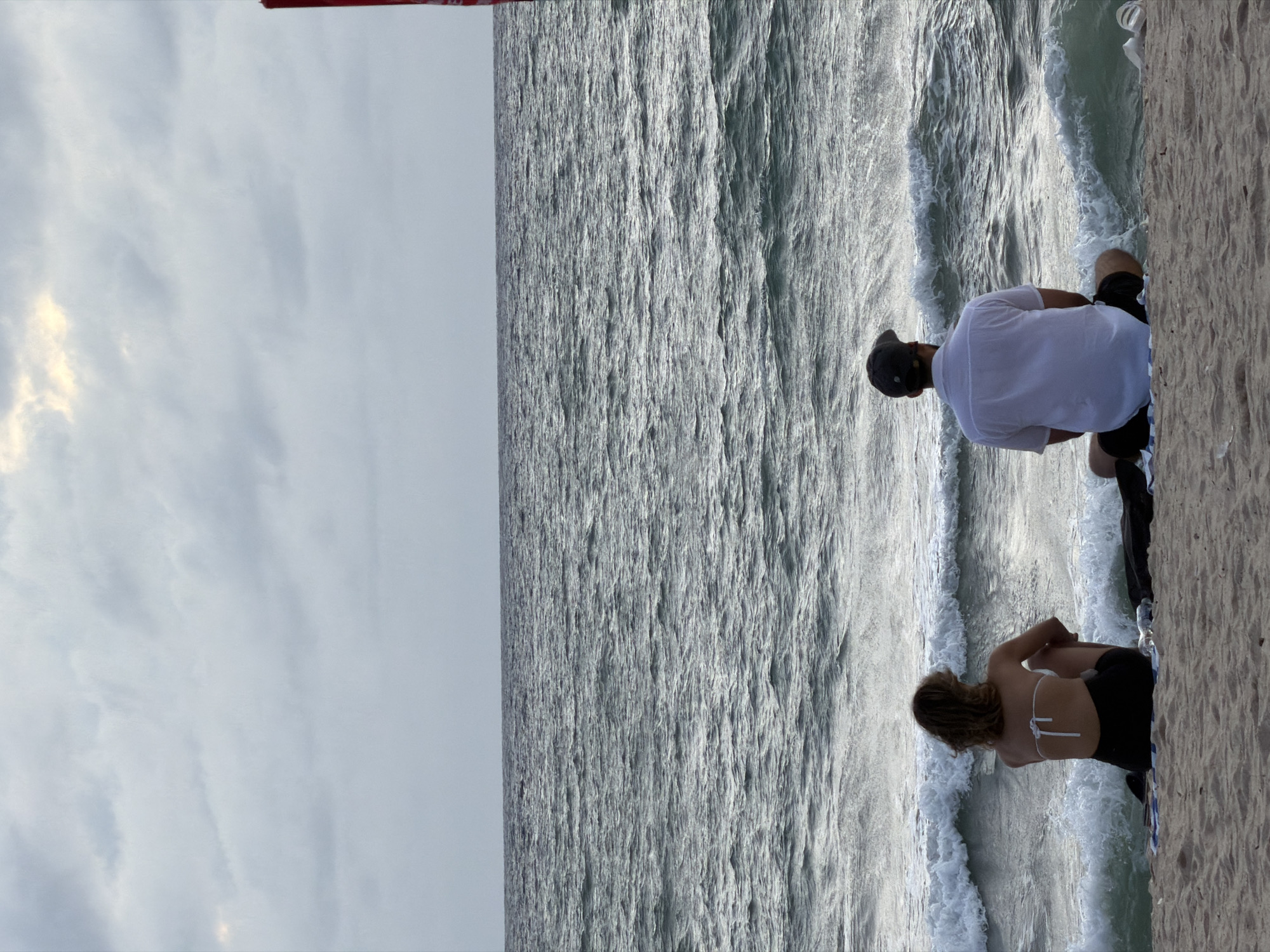 The photograph shows a view from a seawall in Choeng Thale, Thailand. A man and a woman are sitting with their legs dangling over the wall. The man is wearing a white t-shirt, black shorts, and a baseball cap. The woman is wearing a white bikini top and black shorts. They appear to be looking out at the water. Waves are visible, crashing against the wall. The sky is overcast.
