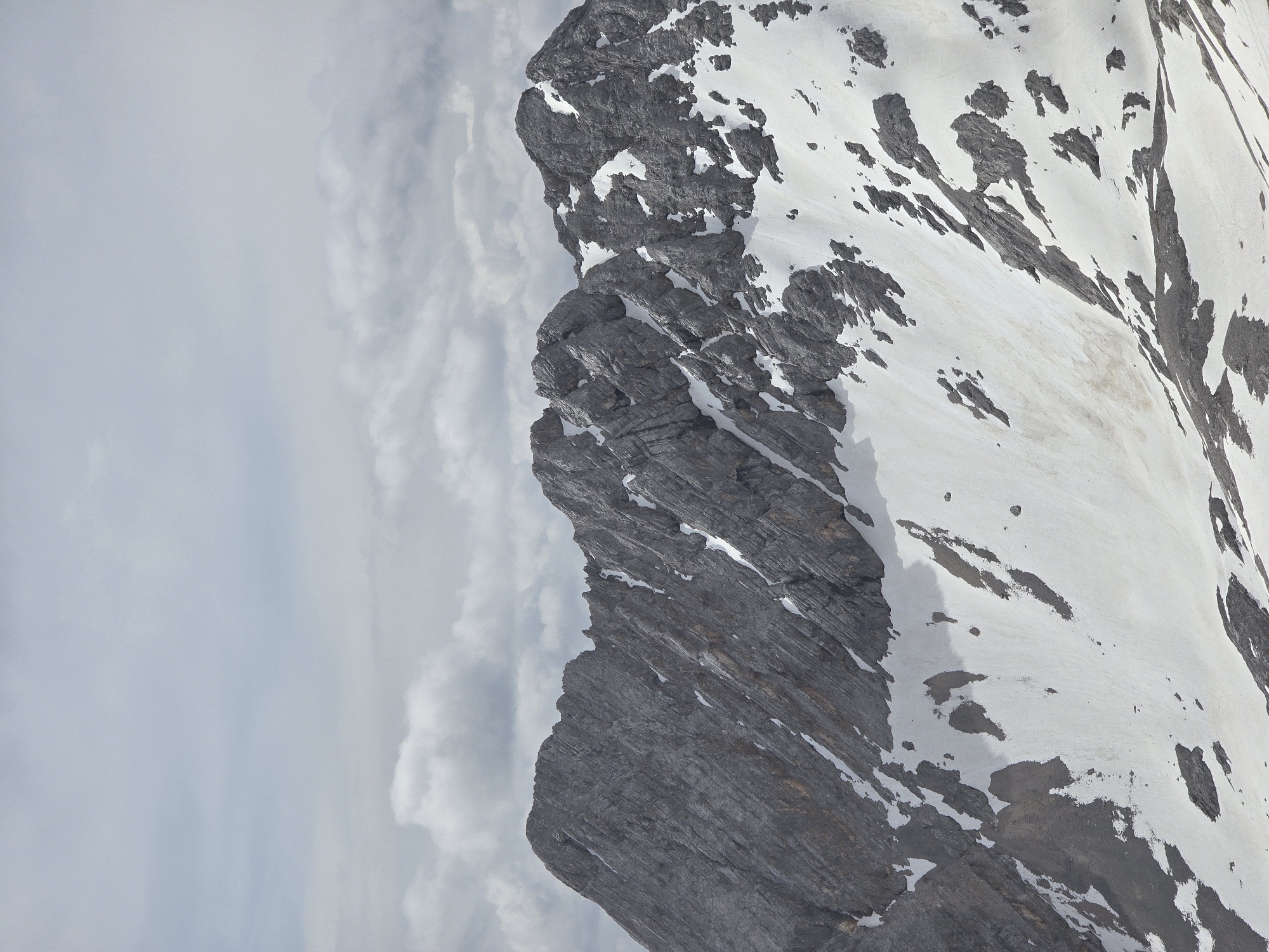 The image shows a mountainous landscape with rocky peaks partially covered in snow. The sky is overcast with clouds. The lower portion of the image shows a slope with patches of snow and exposed grey rock. The upper portion reveals the rugged texture of the mountain ridges. The scene depicts a natural environment, likely at a high altitude. While the image is of a mountainous region, it does not depict Garmisch-Partenkirchen, Germany, or any urban elements.