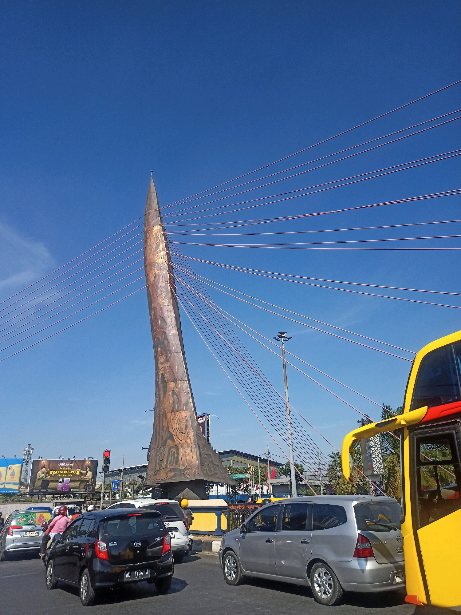 The image shows a busy street scene in Surakarta, Indonesia, dominated by a large, distinctive monument and a cable-stayed bridge. The monument, a tall, slender structure with a textured, metallic surface, stands prominently in the center of the frame, against a clear blue sky. Numerous reddish-brown cables emanate from the top of the monument, stretching outwards and upwards, indicative of the bridge's suspension system.
In the foreground, a traffic jam is evident. Several cars and a motorcycle are stopped on the road. A black hatchback with the license plate "AD 1734 U" is in the center, while a silver minivan is to its right, partially obscured by a bright yellow and red bus on the far right of the image. The bus is a significant portion of the right edge of the frame. A person wearing a red shirt is visible on the motorcycle.
In the background, there are buildings and billboards. One large billboard depicts an advertisement with two people. Traffic lights are visible, with one showing a red light. Palm trees and other greenery can be seen behind the vehicles and monument, suggesting a tropical or subtropical climate. The overall impression is one of urban activity under a bright, sunny sky.