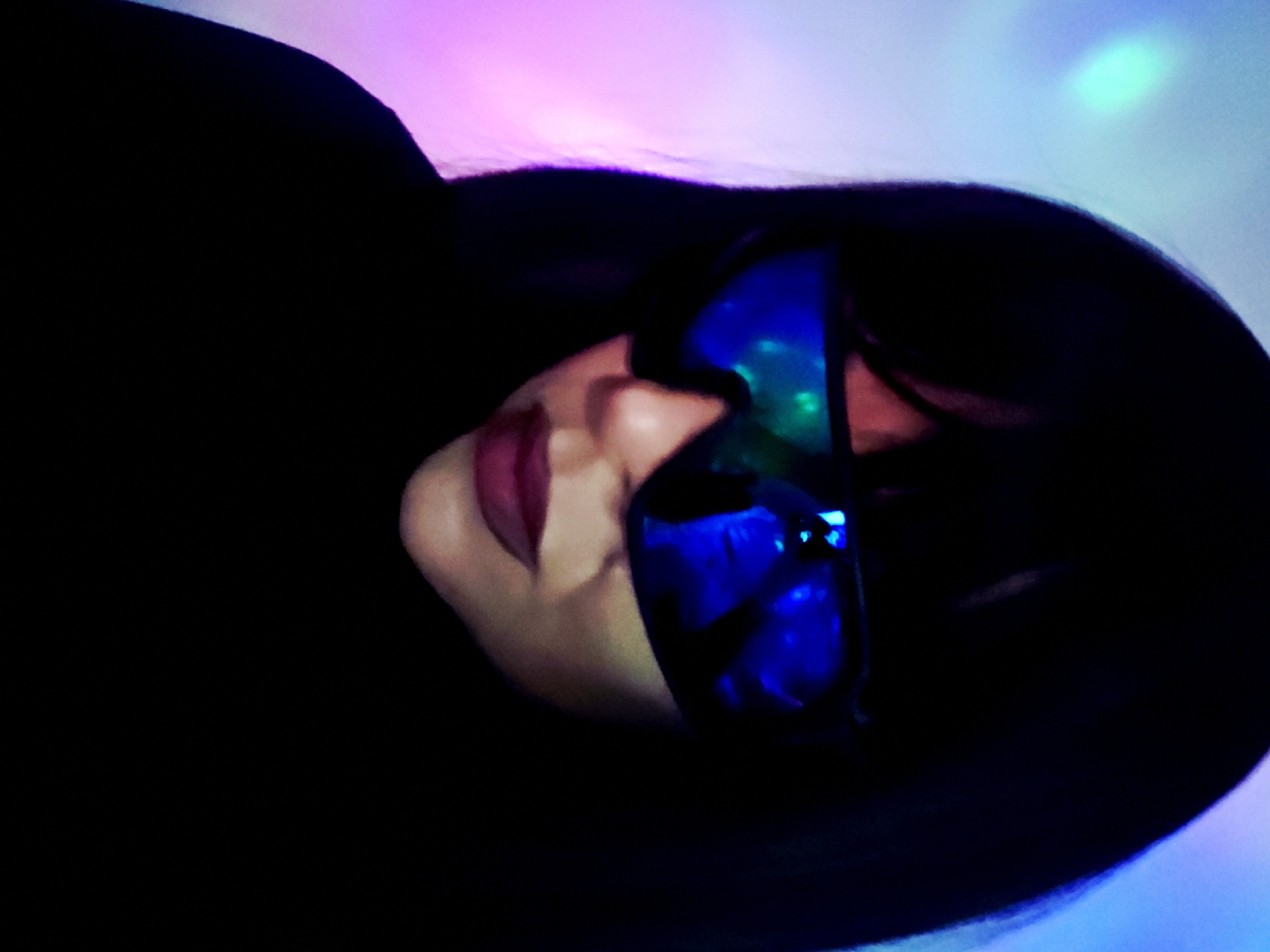 A low-angle, close-up shot features a person's face in dim, club-like lighting with colored ambient light of pink, blue, and green. The person is wearing reflective, blue tinted glasses, covering the eyes. Only the side of the face, mouth with dark red lipstick, and part of the nose are visible. The person's dark hair surrounds their face, largely obscuring the background. The background itself is dark, with scattered, colorful light spots. The location is not possible to determine.