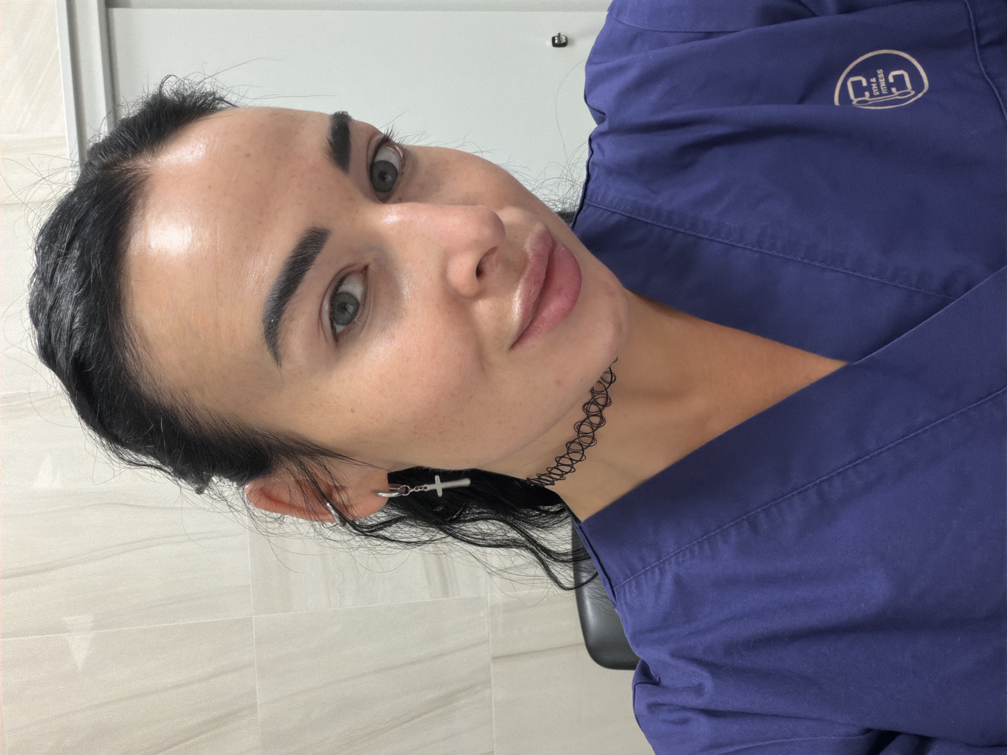A medium close-up shot depicts a fair-skinned woman with dark hair and makeup, wearing a blue collared shirt. The woman is positioned at an angle, facing slightly to the left of the frame. Her hair is pulled back, and she wears dangling cross earrings. A black, patterned choker adorns her neck. The background consists of a neutral-colored wall with tile-like patterns. The letters "GYM & FITNESS" and a stylized logo are printed on the upper right of her shirt. There are no indicators suggesting that the photo was taken in Saint Petersburg, Russia.