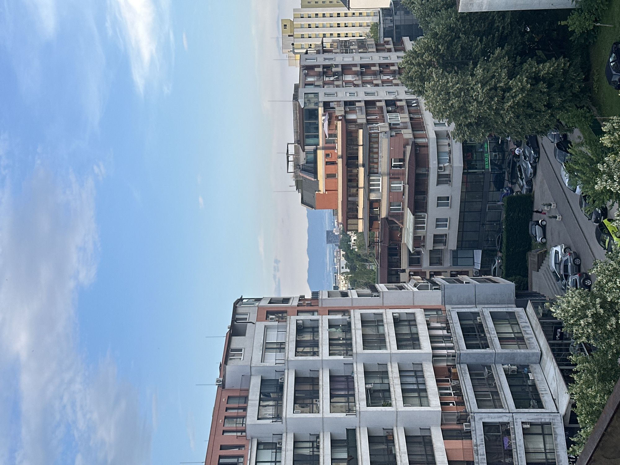 The image shows a view of residential buildings and street life in Sofia, Bulgaria. In the foreground, a modern apartment building with balconies and large windows dominates the left side of the frame. To the right, other apartment buildings of varying architectural styles and heights are visible, including a tall, yellow building in the background. The scene includes parked cars on the street and in a parking lot. Some trees with green foliage are also present, suggesting a pleasant day. In the distance, a hazy cityscape with distant mountains under a blue sky with scattered clouds can be seen. One of the buildings has a sign with Cyrillic lettering that reads "УТОПИЯ". A few people are visible on the sidewalk. The overall impression is that of an urban residential area in Sofia during daylight.