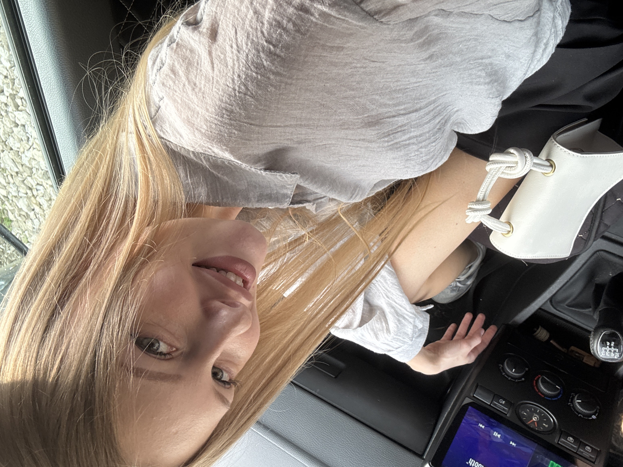 A young Caucasian woman with long blonde hair and green eyes smiles into the camera, taking a selfie inside a car. She is wearing a light gray, crinkled fabric shirt. Her left hand is resting on her leg, and a small white purse with a knotted handle is visible near her right hand. The car's interior, including the dashboard with a digital display and climate controls, is visible. The image is taken from a slightly elevated, close-up perspective, with natural light illuminating the scene. The specific location in the city of Perm, Russia cannot be determined from the visual information alone.