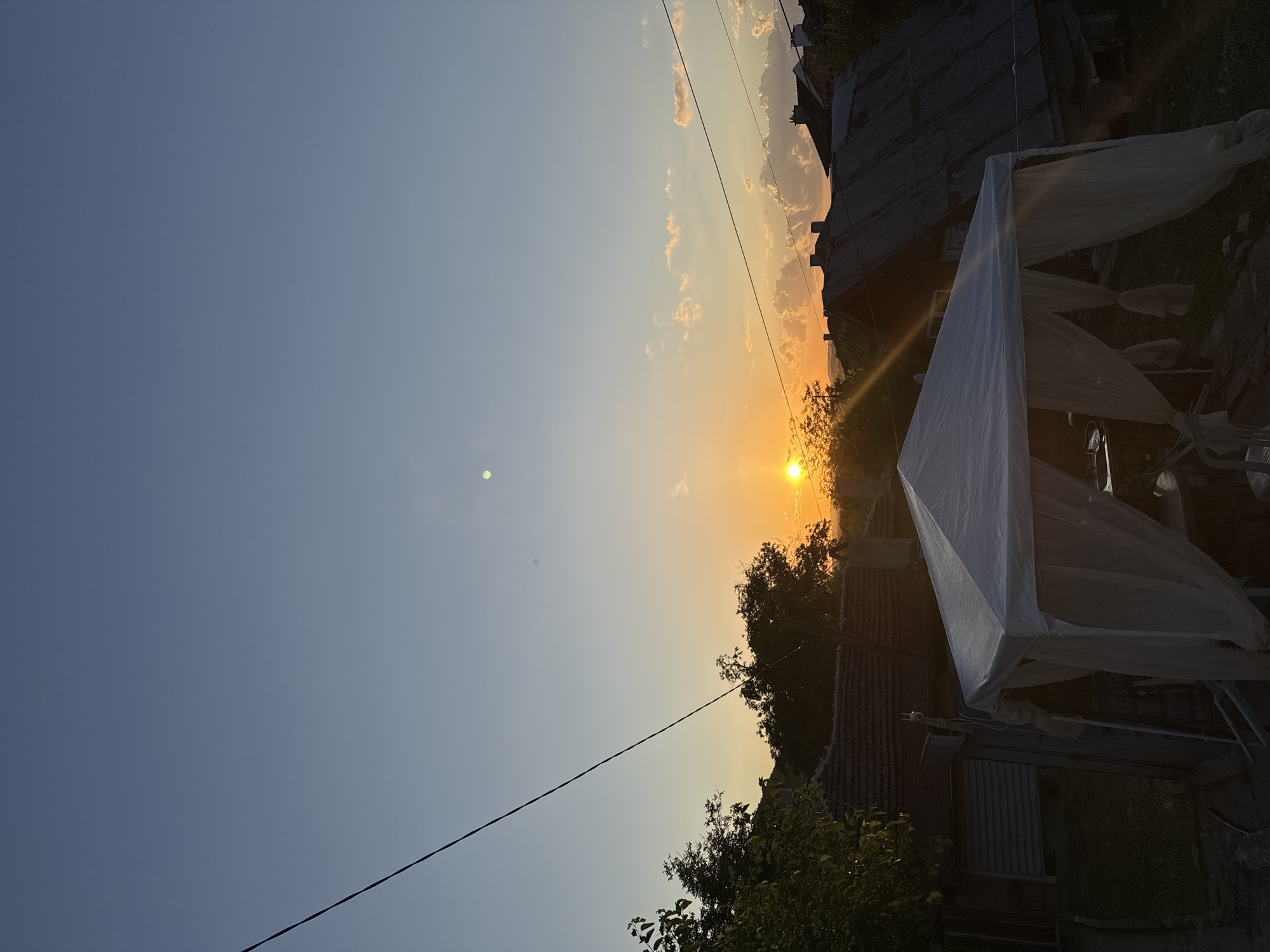 A vertical, full shot captures a rural sunset over Sredets, Bulgaria. The sun, a brilliant orb of orange, hovers just above the horizon, casting a warm glow across the sky, which transitions from a deep blue at the top to hues of orange and yellow near the setting sun. Wispy clouds are scattered across the sky, some catching the golden light. In the foreground, a white canopy tent with sheer white curtains is set up, partially obscuring a dark yard. Behind the tent, the rooftops of several houses are visible, some appearing weathered. Trees with lush green foliage flank the scene, silhouetted against the vibrant sky. Power lines are visible crisscrossing the upper portion of the image. The overall impression is one of a peaceful evening in a suburban or rural setting.
