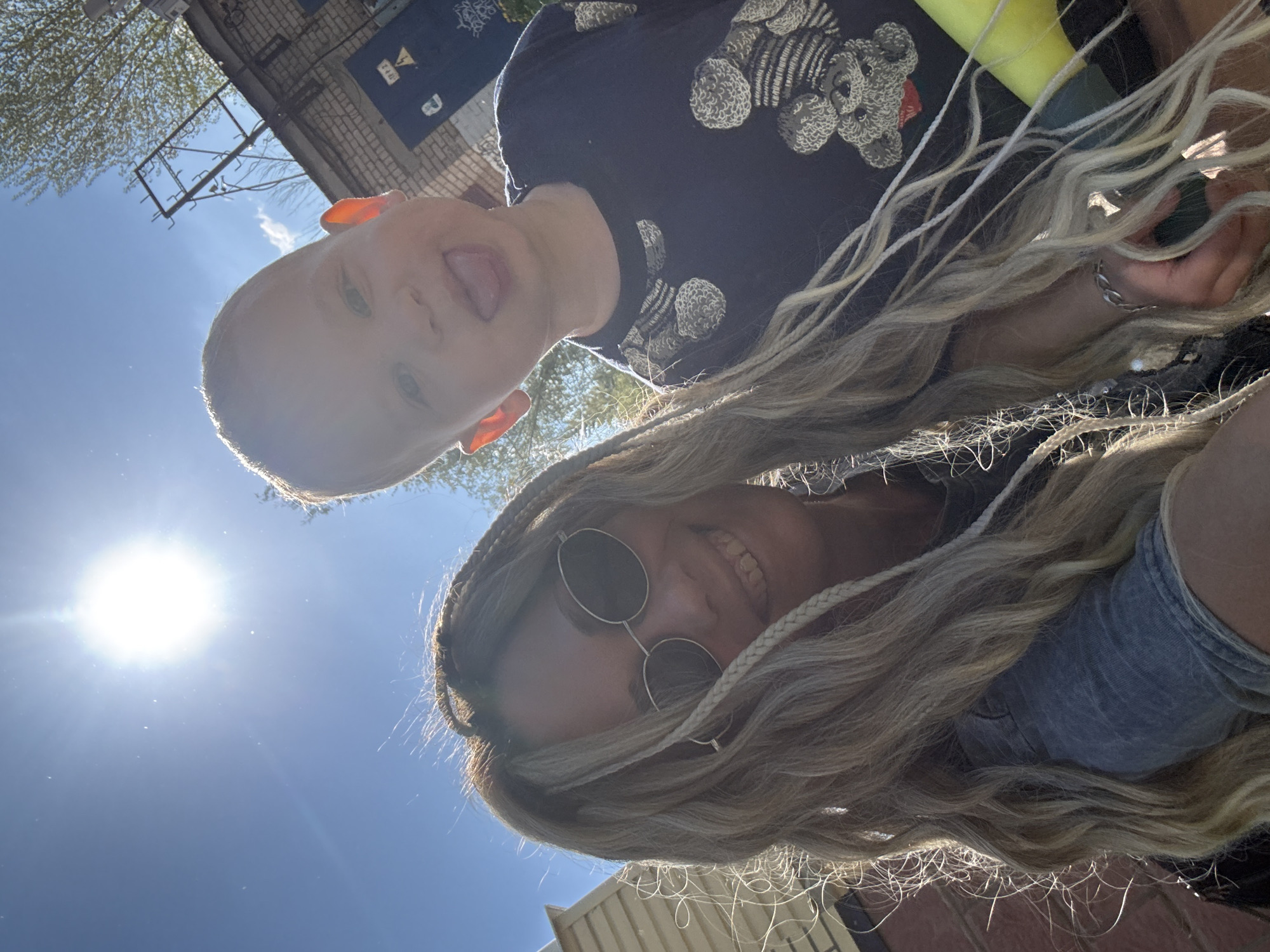 A low-angle selfie shows a young child and adult, possibly a parent, outdoors. The child, with fair skin and light hair, sticks their tongue out towards the camera. They are wearing a dark-colored shirt with a teddy bear pattern. The adult, wearing round sunglasses, has blonde hair with braids and a blue denim shirt. The background shows a blue sky and building exteriors. The location is identified as Rusanovo, Russia.