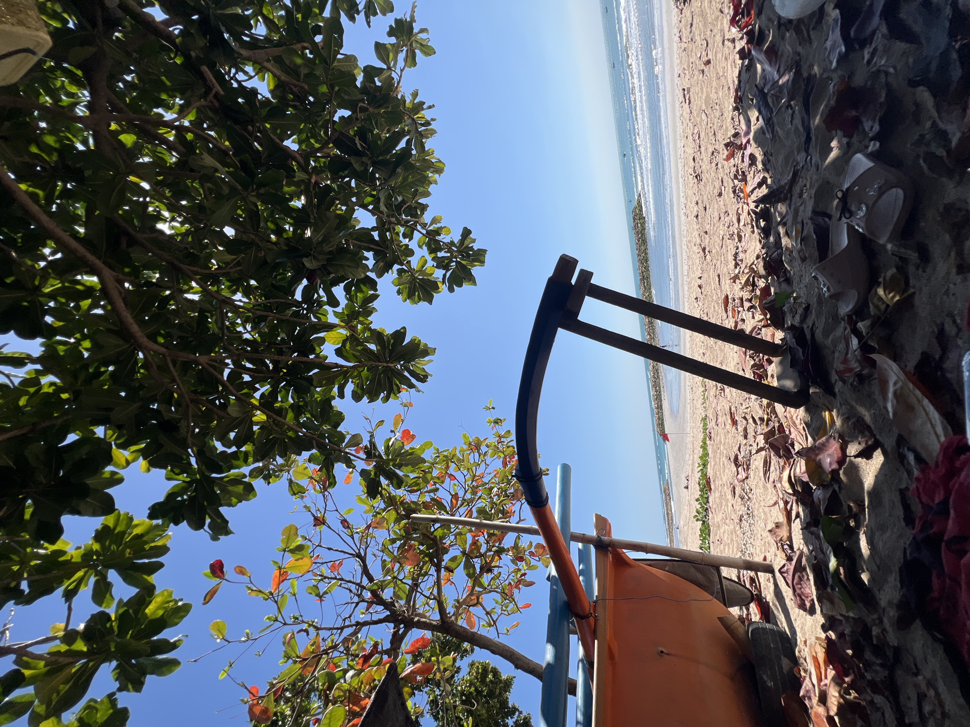 This image depicts a tranquil beach scene in Kuta, Indonesia, likely during daylight hours with a clear blue sky. The foreground is covered in dried leaves and sand, with a pair of light-colored sandals visible on the right. A portion of an orange boat is present on the left, supported by wooden and metal structures. In the midground, the sandy beach stretches towards the ocean, with gentle waves lapping the shore. A stone breakwater is visible further out, with a few small boats dotting the water. A red flag is planted on the beach in the distance. The background is dominated by the expansive ocean and the clear sky, with lush green foliage from trees overhead casting shadows on the scene. The overall impression is one of a peaceful, natural setting.