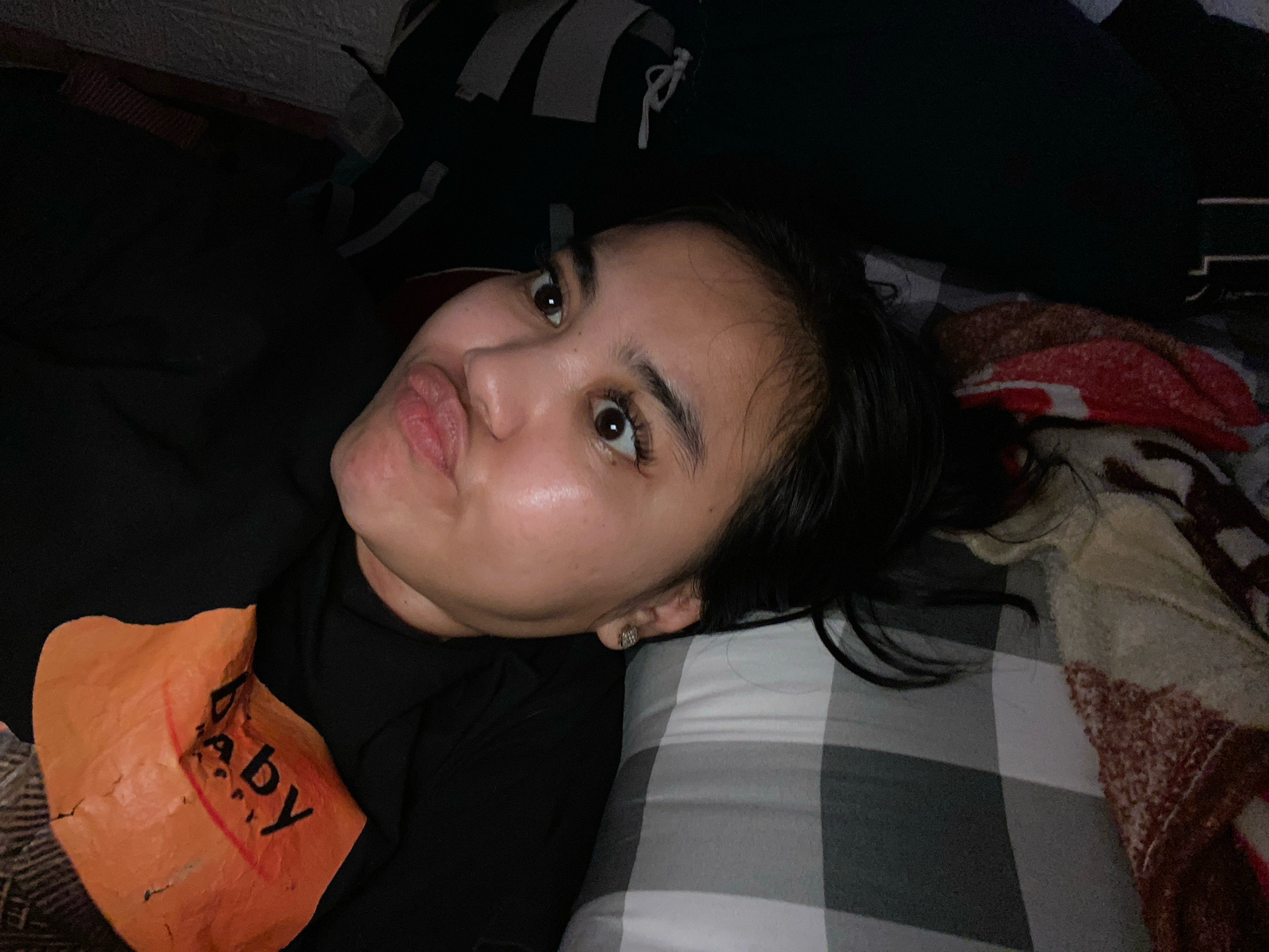 A young woman with dark hair and light skin lies on her side, looking at the camera. She has her lips pursed in a "duck face" pose. She is wearing a black t-shirt with an orange graphic on the front that appears to say "Baby". She has a small hoop earring in her left ear. The background consists of bedding with a plaid pattern. The scene appears to be indoors, likely in a bedroom. The lighting is dim. The location is the city of Hegarmukti, Indonesia.
