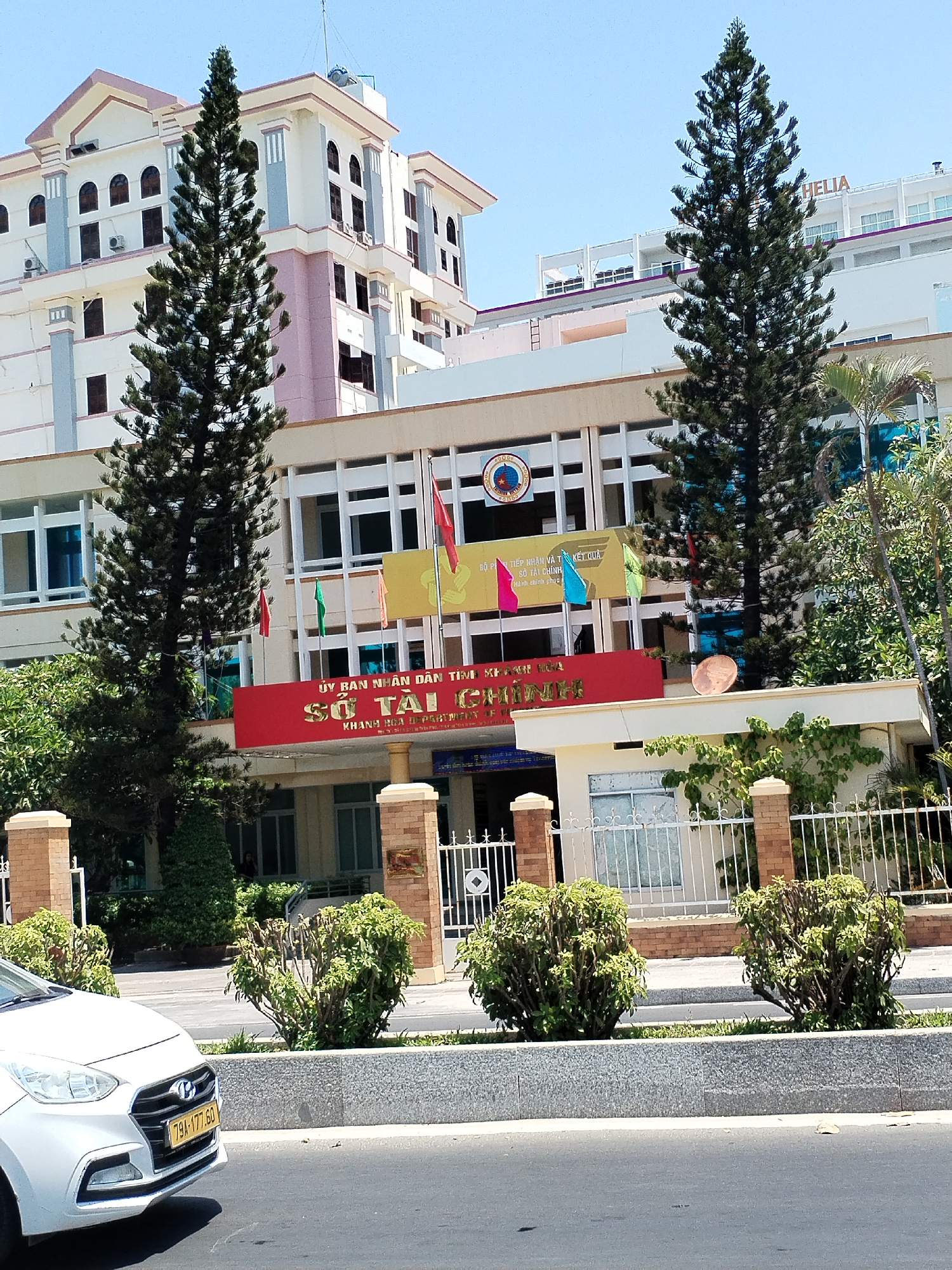 The image depicts a building identified as the "Department of Finance" of Khanh Hoa province in Nha Trang, Vietnam. The building features a red sign reading "Sở Tài Chính," above which is a yellow banner with Vietnamese script. Several flags are visible on top of the building. Two tall, thin evergreen trees flank the building's façade. A white car is partially visible in the foreground on the left. There's a gated fence in front of the building, with trimmed bushes lining the base of the fence. In the background, there are other buildings, including one with the name "Helia" on its roof. The sky is clear.