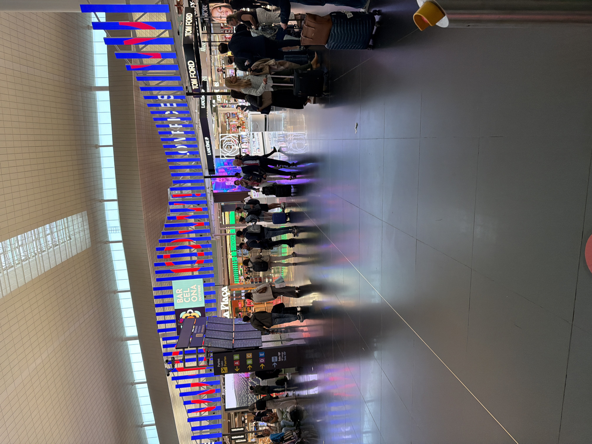 This indoor shot depicts what appears to be a concourse in an airport, possibly in El Prat de Llobregat, Spain. A line of people, possibly passengers, is standing in the center, facing towards the right. Many are pulling luggage. Above the line is a sequence of store signs for luxury brands, including TOM FORD, DIOR, and LANCOME. On the left, signs indicate "BARCELONA DUTYFREE", gates, and VIP areas. A blue and red decorative element runs along the ceiling. The floor is dark grey, reflecting the scene. Light fixtures are visible in the upper-right corner. The shot appears to be taken at an angle, creating a distorted perspective of the scene.
