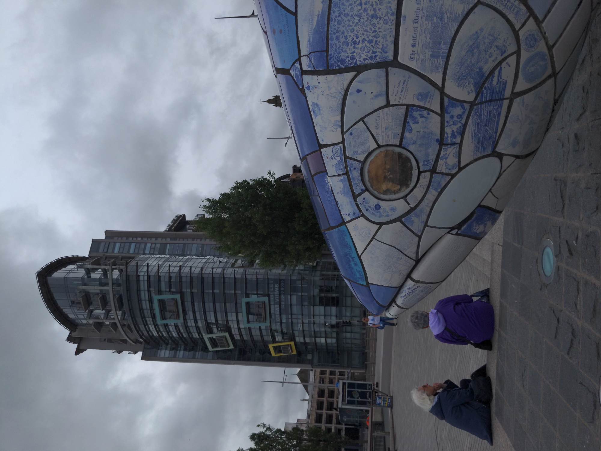 The image captures an outdoor scene on a cloudy day, likely in Belfast, United Kingdom. In the foreground, a large mosaic sculpture of a fish head, primarily blue and white, dominates the right side of the frame. The mosaic features various patterns and text fragments.
In the middle ground, two individuals are seated on a paved area. The person on the left, with white hair and wearing glasses, is dressed in a dark jacket and appears to be looking towards the right. The person on the right has grey hair, is wearing a purple hoodie, and is facing away from the camera.
Behind them, a modern, tall building with a curved glass facade and balconies stands prominently. The building is dark grey and reflects the overcast sky. Signs are visible on the building, including one with the text "WITH GREAT VIEWS LET" and a phone number. To the left of this building, a partially visible structure with rows of parking spaces can be seen.
The ground is paved with dark, textured stones. A small, circular light fixture is embedded in the pavement in the lower foreground. The overall environment suggests an urban public space.