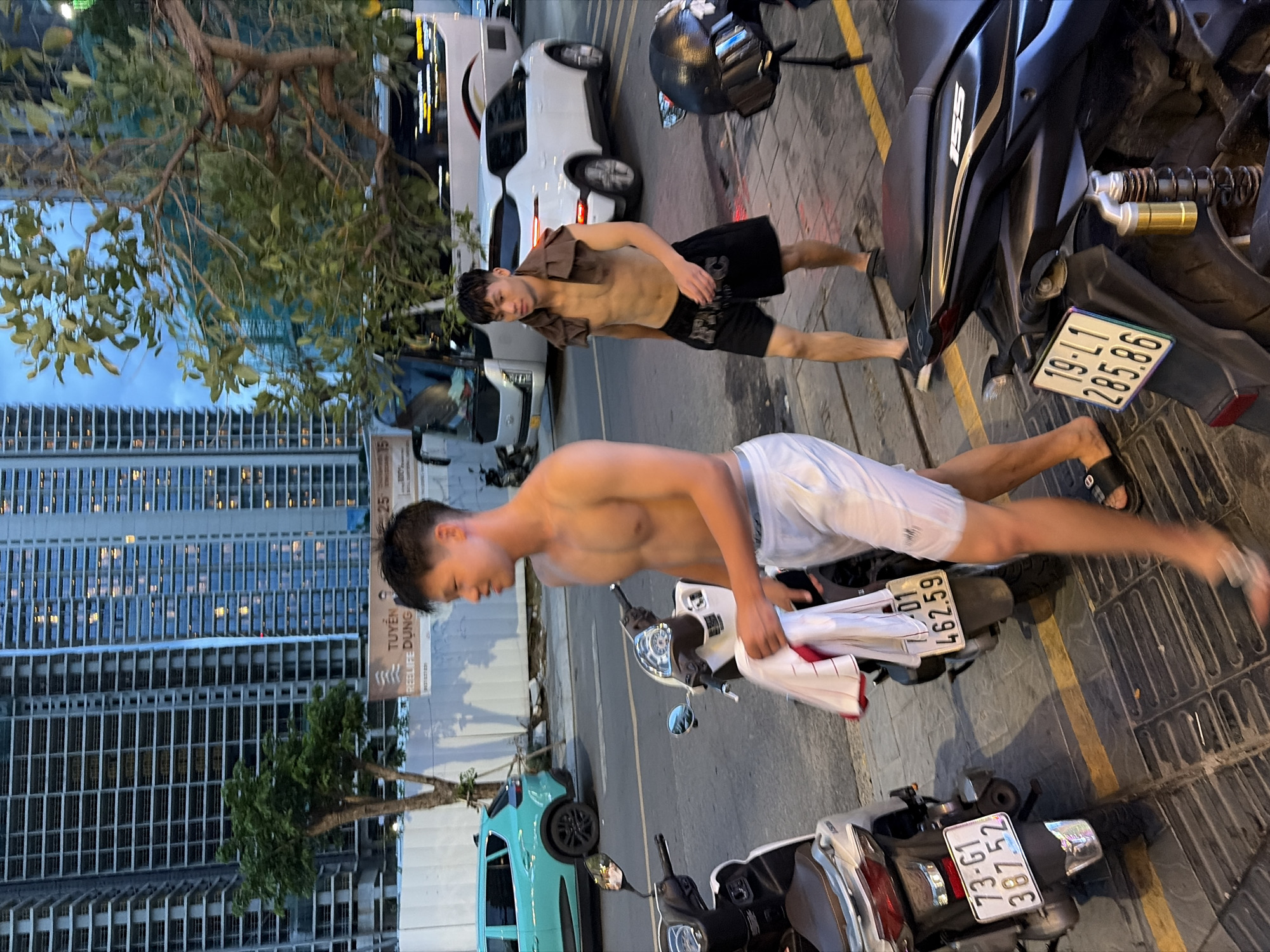 The image depicts two shirtless young men walking on a sidewalk in Đà Nẵng, Vietnam, with a backdrop of high-rise buildings under construction and a street with parked motorcycles and vehicles. The man in the foreground, wearing white shorts, is carrying a bundle of clothing. The man in the background is wearing black shorts and has a towel draped over his shoulders. Several motorcycles are visible, including their license plates. A turquoise car is parked on the street, and a white SUV is driving in the background. The sky is overcast, suggesting it might be late afternoon or evening. The scene appears to be a busy urban environment.