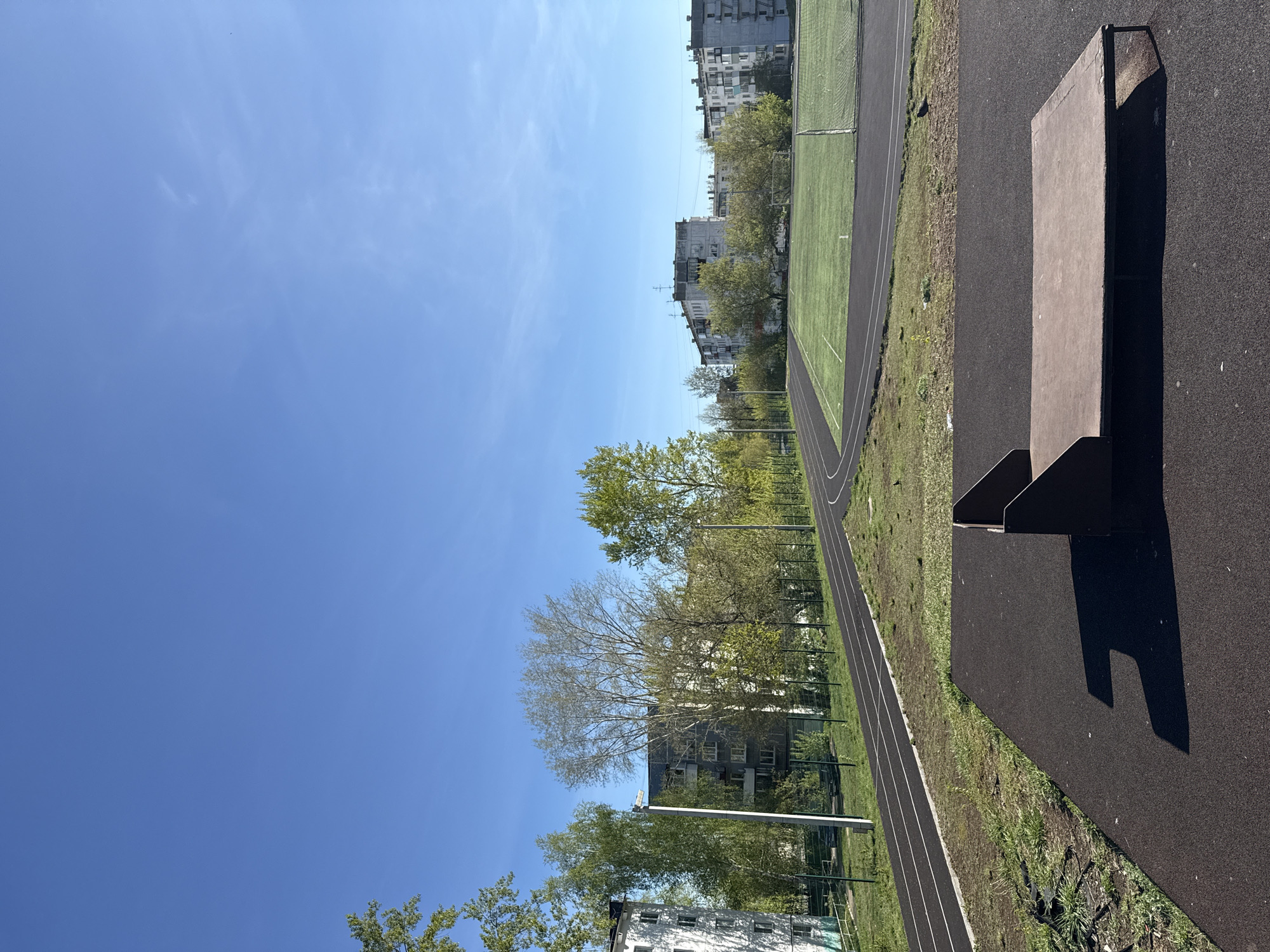 The image depicts a track and field area in the city of Chaikovsky, Russia. The scene includes a running track, a soccer field, and a long jump apparatus. There are apartment buildings visible in the background, along with trees along the edge of the field. The sky is clear and blue.