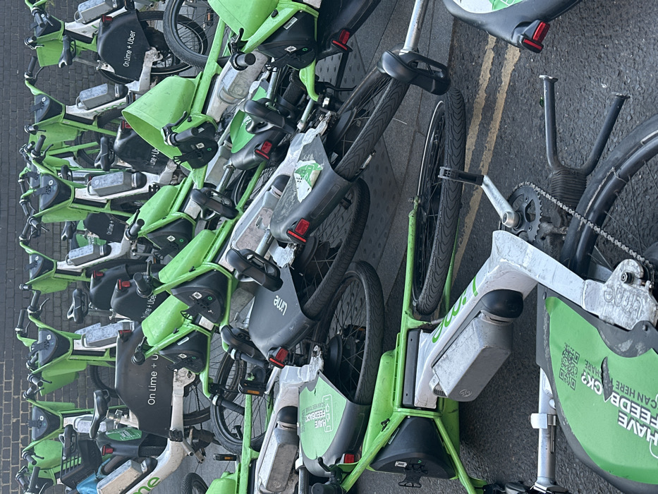 The scene depicts a large accumulation of electric bicycles, predominantly identifiable as 'Lime' bikes by their bright green frames and black components, in a state of disarray on an urban street in London, United Kingdom. Many bikes are lying horizontally on an asphalt surface, some stacked on top of others, forming a chaotic pile that dominates the foreground and midground. In the background, a denser cluster of Lime bikes, some upright, stands against a dark brick wall. 'On Lime + Uber' branding is visible on several bike frames. The ground features double yellow lines on the asphalt, consistent with UK road markings, adjacent to textured grey pavement. No individuals are present.