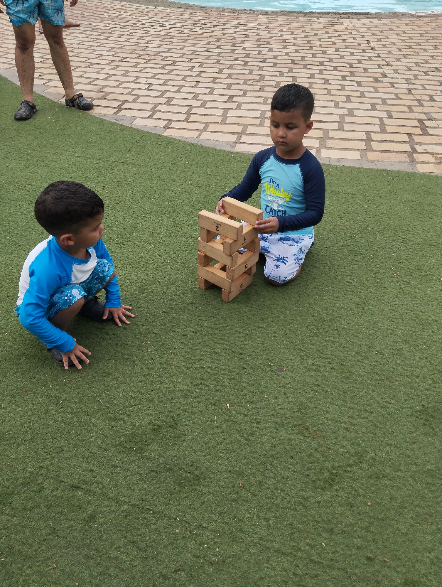 Two young boys are playing with wooden blocks on a green surface next to a brick patio in Porlamar, Venezuela. The boy on the left is crouching and looking at the other boy, who is kneeling and placing a block on a tower. The boy on the left wears a blue and white swimsuit with long sleeves, and the boy on the right wears a similar blue swimsuit. A person's legs are visible in the upper left corner of the image. A pool is visible in the background.