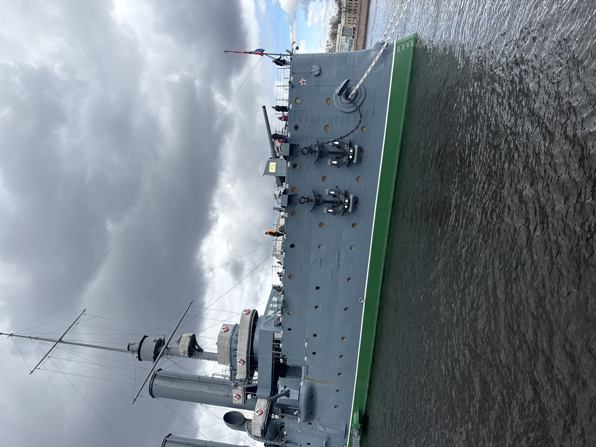 The image shows the Russian protected cruiser Aurora, a museum ship docked in Saint Petersburg, Russia. The ship is painted in gray with a green waterline and features a number of large guns, anchors, and a tall mast. Several people are visible on the deck. The water alongside the ship is dark and choppy. The sky is overcast with heavy clouds. Buildings are visible in the background.
