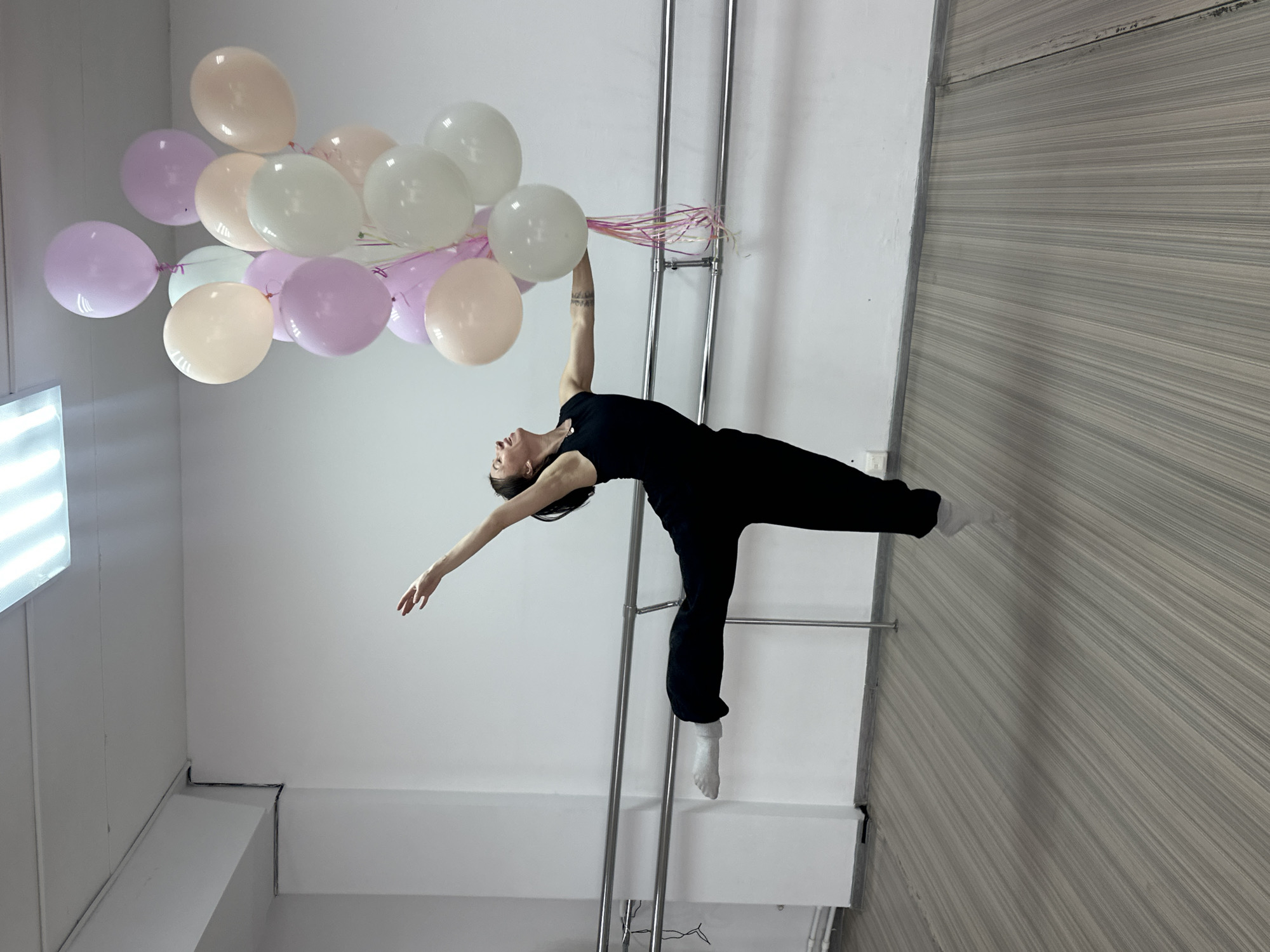 A woman in black clothing and white socks is performing a dance move in an indoor space. She is holding a bunch of pastel-colored balloons (lavender, peach, and white) with her right hand, while her left arm is extended. The woman is positioned sideways, balancing with one foot on a metal structure that appears to be a ladder or ballet barre. The space has white walls and a wall with striped wallpaper. Based on the metadata, the location is Votkinsk, Russia.