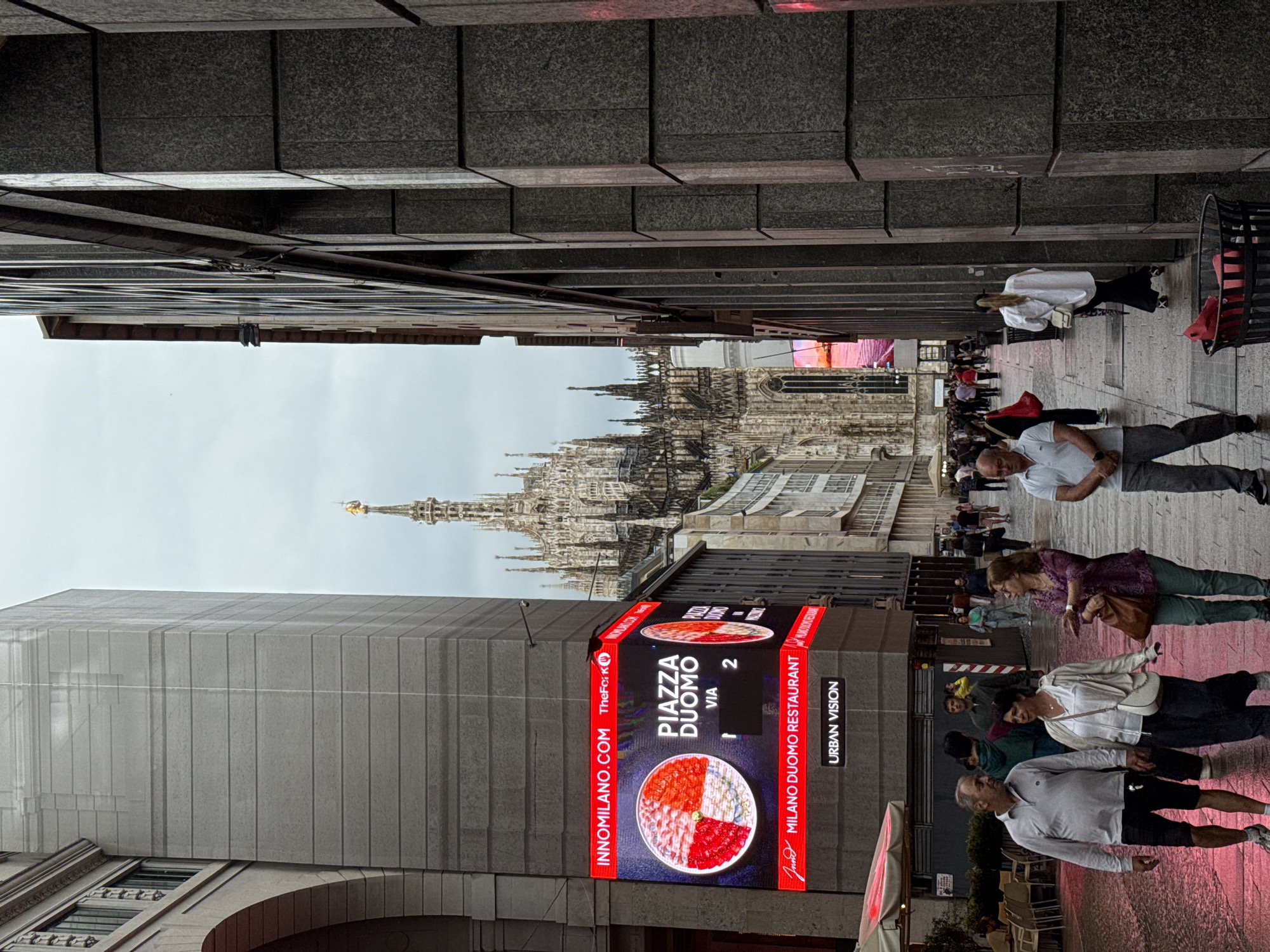 The image depicts a bustling urban street or pedestrian thoroughfare in Milan, Italy, with the iconic Milan Duomo visible in the distant background. Tall, modern-looking buildings with grey paneled facades line both sides of the street.
On the left, a prominent digital billboard, largely red with black accents, advertises a restaurant. It displays an image of a multi-sectioned dish, resembling a pizza or similar Italian appetizer. Visible text on the billboard includes "INNOMILANO.COM", "TheFork", "PIAZZA DUOMO VIA 2", "MILANO DUOMO RESTAURANT", and "URBAN VISION".
Numerous pedestrians are walking along the street. In the foreground on the right, a man in a white polo shirt and dark shorts walks towards the viewer. Further back, a woman in a white top and dark pants, carrying a white handbag, is also walking. Another woman in a purple blouse and green trousers, holding a brown bag, is seen walking towards the viewer on the right side. Other individuals and small groups are scattered throughout the midground, engaged in walking or standing.
The magnificent Gothic spires of the Milan Duomo dominate the distant background, rising above surrounding buildings under an overcast, light grey sky. The golden statue atop its highest spire is clearly distinguishable. The scene suggests a daytime setting, likely a popular area given the concentration of people and the landmark in view. The atmosphere appears to be that of a typical busy city day.