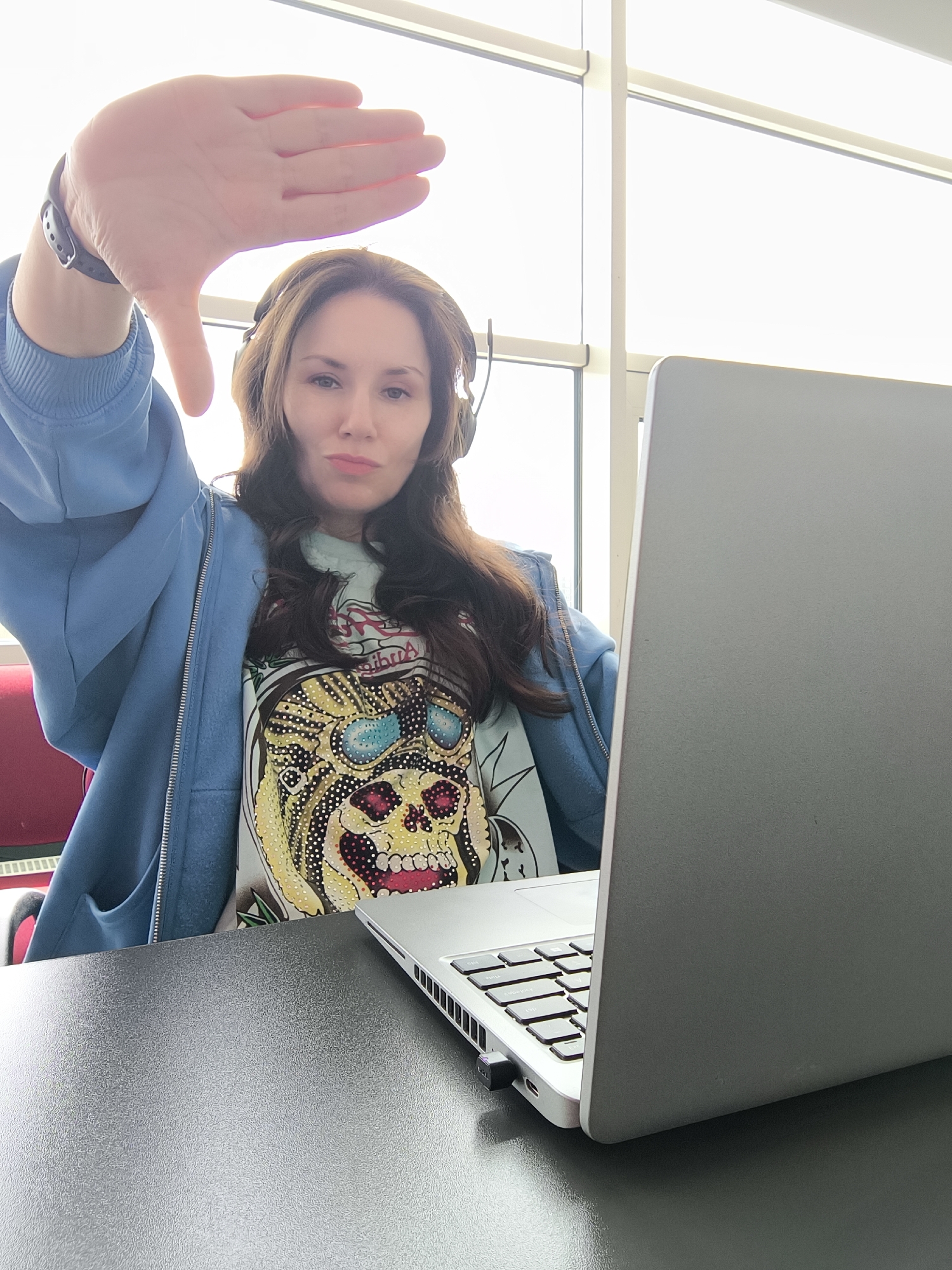 A woman with long brown hair is seated at a black table in front of a laptop computer. She is wearing a blue zip-up hoodie over a light-colored t-shirt with a skull design. She has headphones with a microphone headset on. She is holding her left hand up with her thumb pointing downwards in a "thumbs down" gesture. Behind her, there is a large window with white frames. The location is in a city, though the name is Unknown.