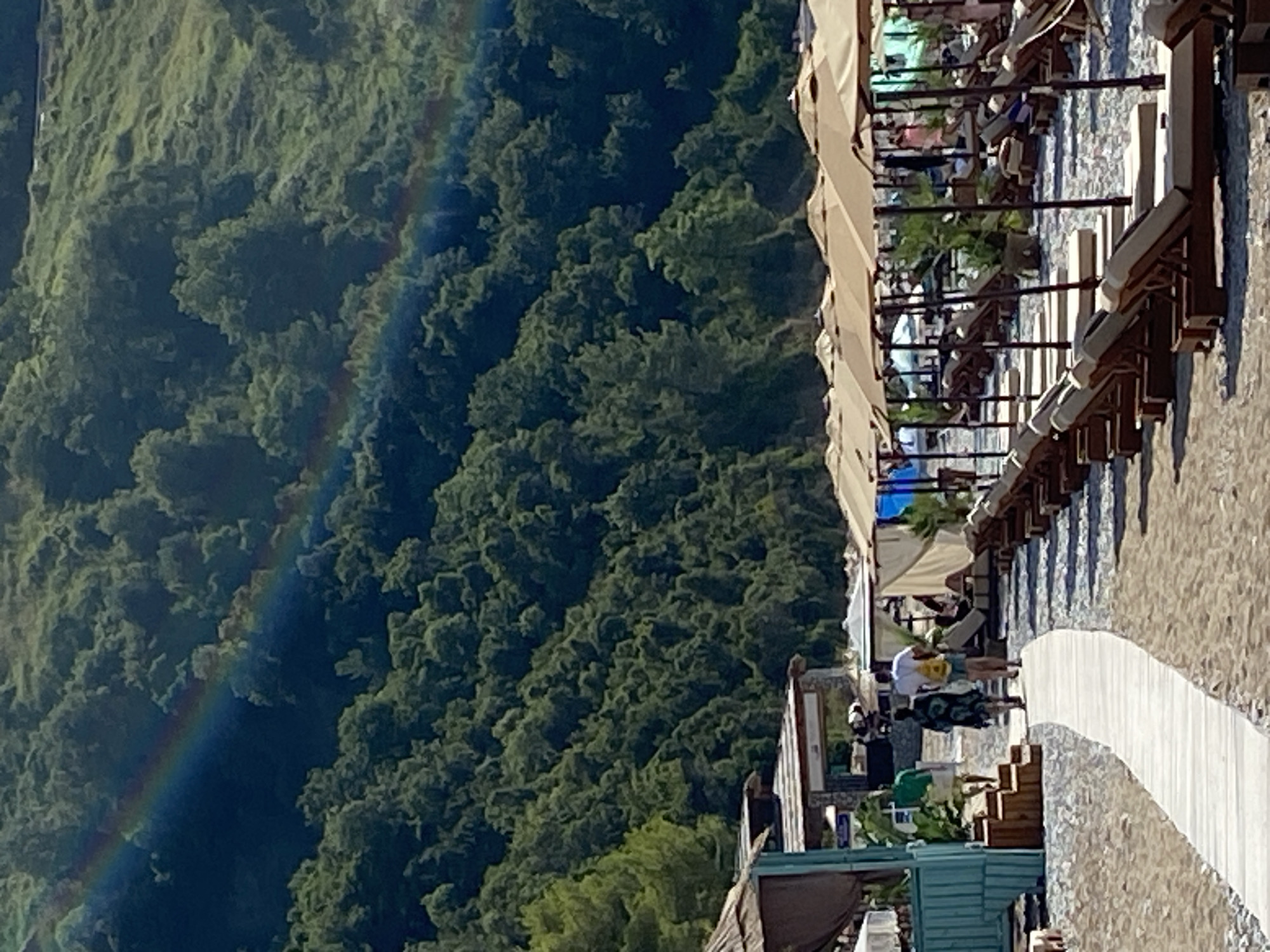 The image depicts a beach scene in Prijevor, Montenegro, characterized by a lush, tree-covered hillside dominating the background. A faint rainbow arcs across the upper portion of the hillside. In the foreground, a sandy beach is lined with rows of sun loungers and umbrellas, many of which are tan in color. A wooden walkway extends into the scene, with three individuals walking away from the viewer on it. One person wears a green shirt, another a patterned blue and black dress, and the third a white shirt and shorts with a yellow backpack. To the left, a teal-colored structure with a thatched roof is visible. The overall atmosphere suggests a sunny day at a resort or beach area.