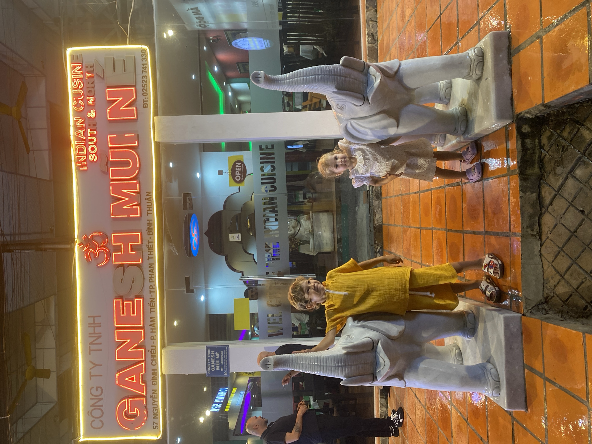 The image shows the exterior of an Indian restaurant named Ganesh Mui Ne, located in Mũi Né, Vietnam. The restaurant features a large, illuminated sign in red neon lettering that reads "CÔNG TY TNHH GANESH MUI NE" on the left and "INDIAN CUISINE SOUTH & NORTH" and "MUI NE" on the right. Below the main sign, smaller text indicates the address as "57 NGUYỄN ĐÌNH CHIỂU - P. HÀM TIẾN - TP. PHAN THIẾT - BÌNH THUẬN" and a phone number "DT: 02523 741 330".
In the foreground, two children are posing with two white elephant statues. A young boy, wearing a yellow outfit, stands to the left of the boy and is touching the trunk of an elephant statue. To the right, a young girl in a light-colored dress stands next to another elephant statue. The ground is paved with orange tiles, which appear wet, suggesting recent rain or cleaning.
Behind the children and elephant statues, the glass facade of the restaurant is visible. Through the glass, interior lighting and restaurant seating can be seen, along with a few patrons. A sign that reads "OPEN 10AM - 10PM" is also visible on the glass door. On the far left, a man in dark clothing stands near the entrance. The overall scene captures a moment outside a restaurant in Mũi Né.