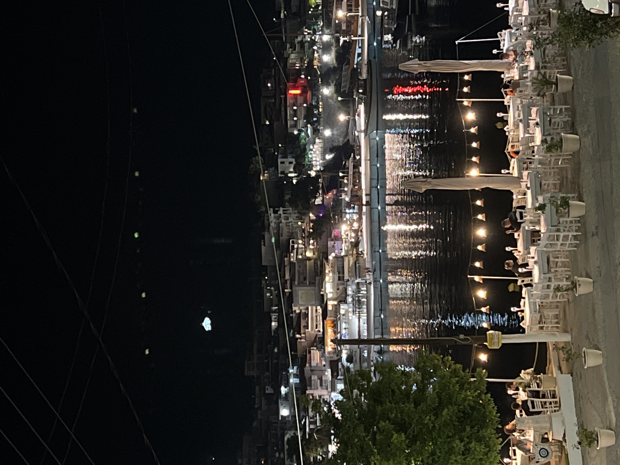 This image depicts a nighttime scene at a waterfront restaurant in Sithonia, Greece. White tables and chairs are set up outdoors, illuminated by strings of lights and reflections from the water. Several people are seated at the tables, appearing to be dining and socializing. Beyond the restaurant, a town with illuminated buildings stretches along the coastline, and boats are docked in the harbor. The water is dark, reflecting the lights of the town and the overhead string lights. The overall atmosphere suggests a lively evening at a seaside establishment.