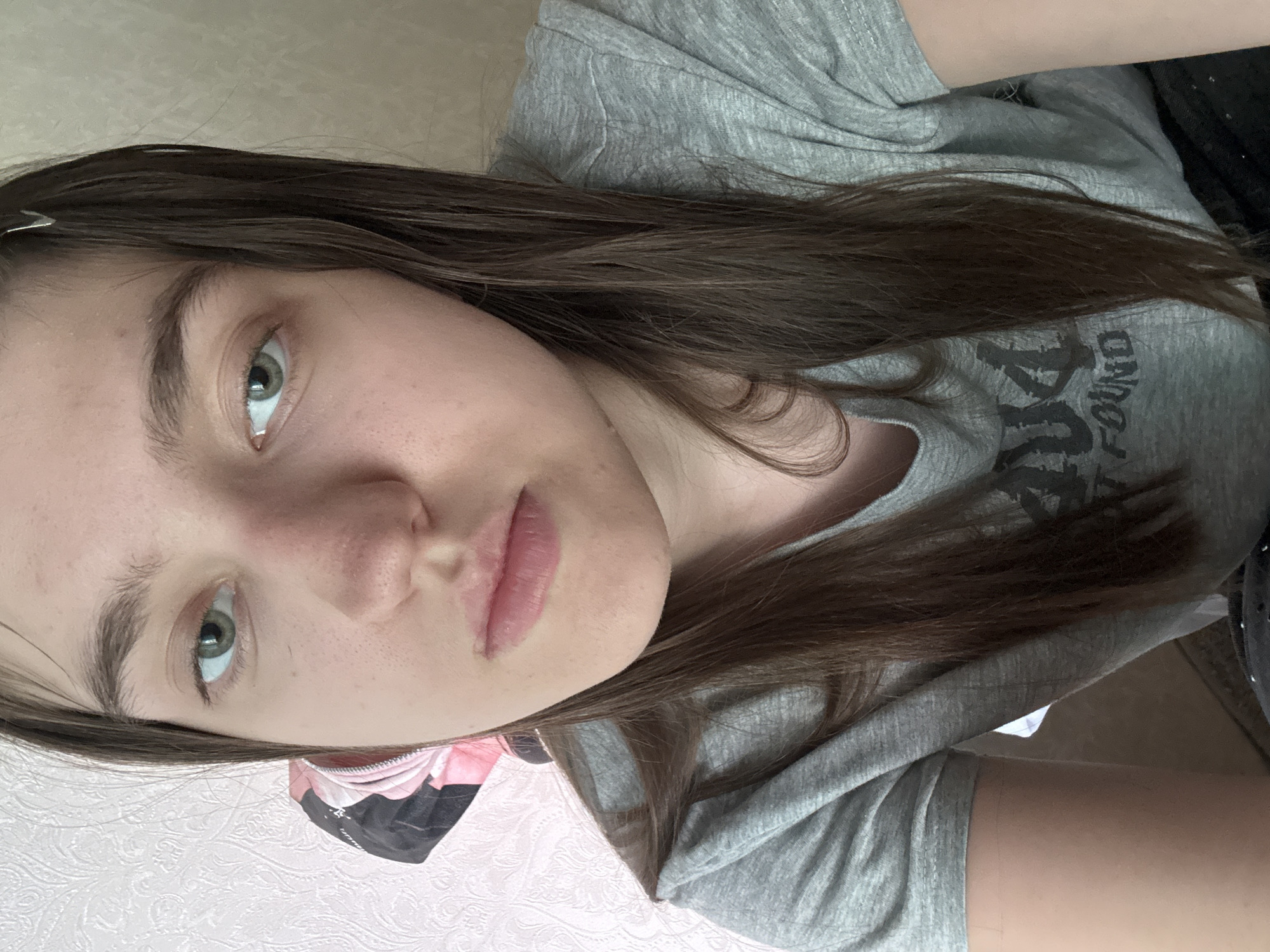 A close-up, slightly tilted portrait captures a young woman with fair skin, green eyes, and light brown hair. A silver hair clip is visible in her hair. She is wearing a grey t-shirt with a faint graphic print. Part of a black and pink object is visible to the left of her face, likely a mask. The background features a textured white wall. The location appears to be indoors. There is no definitive information about the city.