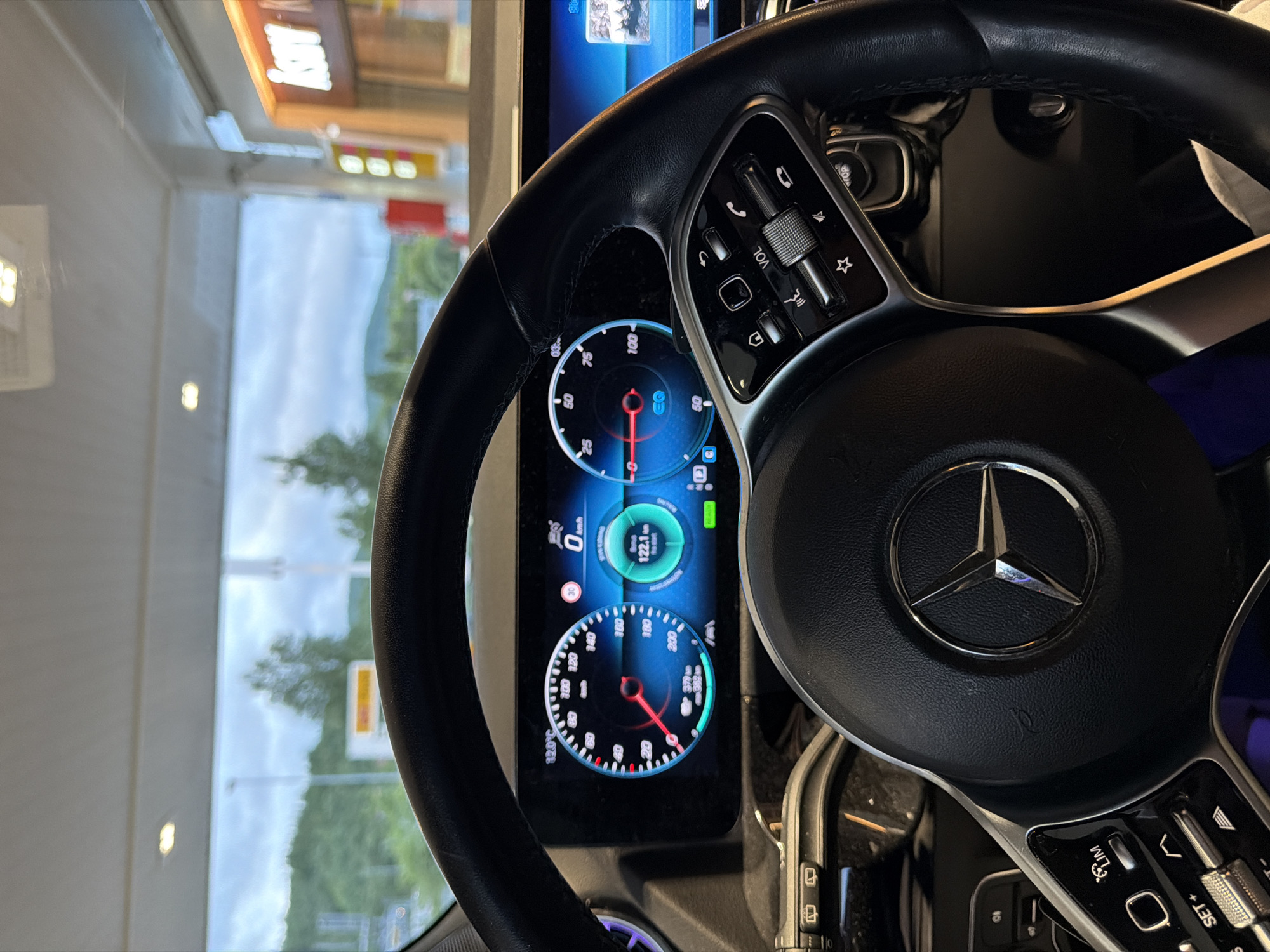 The image shows the interior of a Mercedes-Benz car, specifically the dashboard and steering wheel, with a view of the exterior through the windshield. The dashboard displays a digital speedometer, tachometer, and a trip computer showing a speed of 0 km/h and a distance of 122.1 km. The car appears to be parked at a gas station, indicated by the presence of gas pumps and signage visible through the windshield, including what appears to be a Shell logo. The weather outside is overcast with green hills in the background. The location is identified as Harstad, Norway.