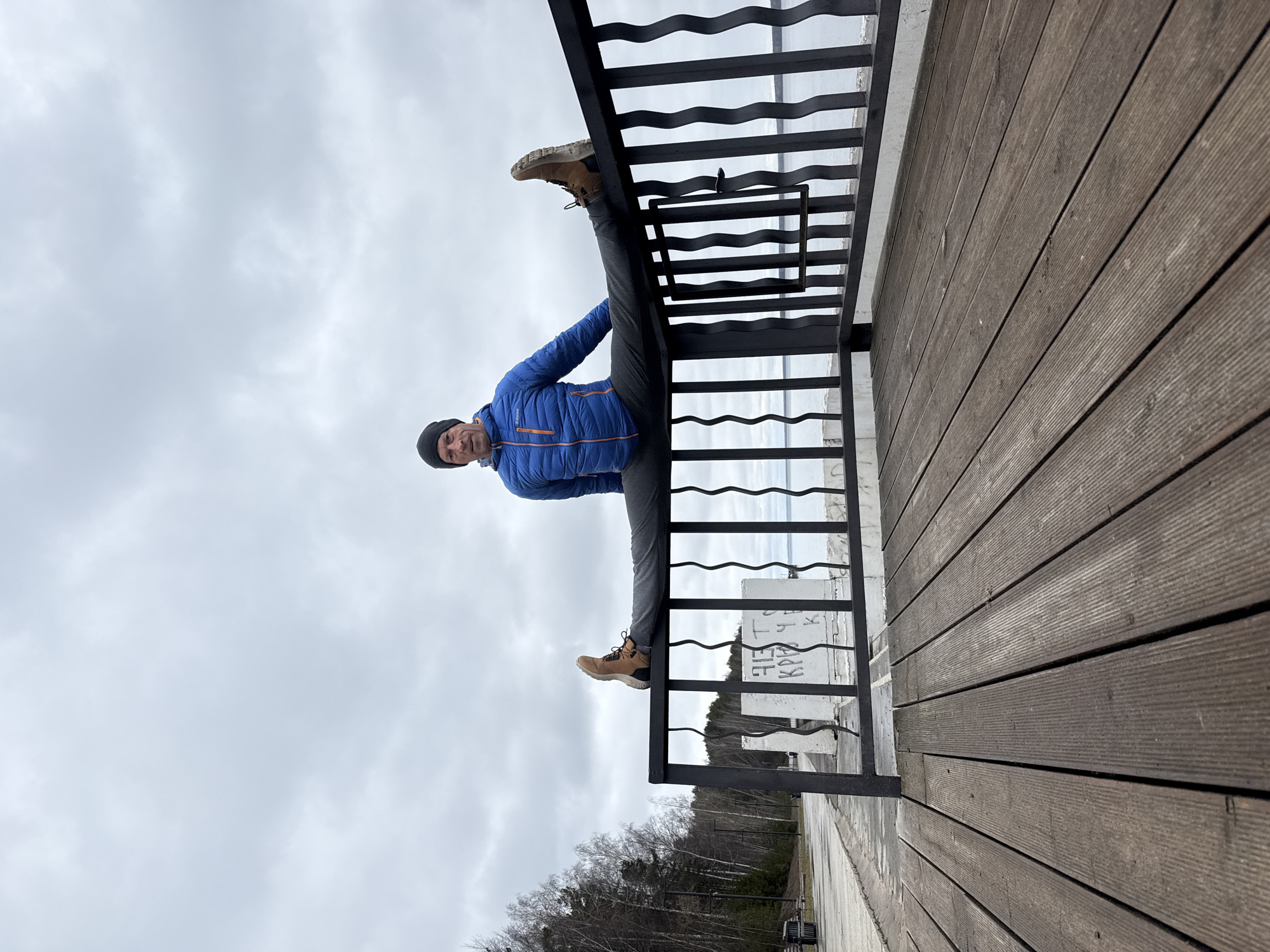 A man performs a split on a metal railing on a wooden deck in Chaikovsky, Russia. The man wears a blue jacket, gray pants, brown boots, and a black hat. The background shows a cloudy sky, trees, and a road.