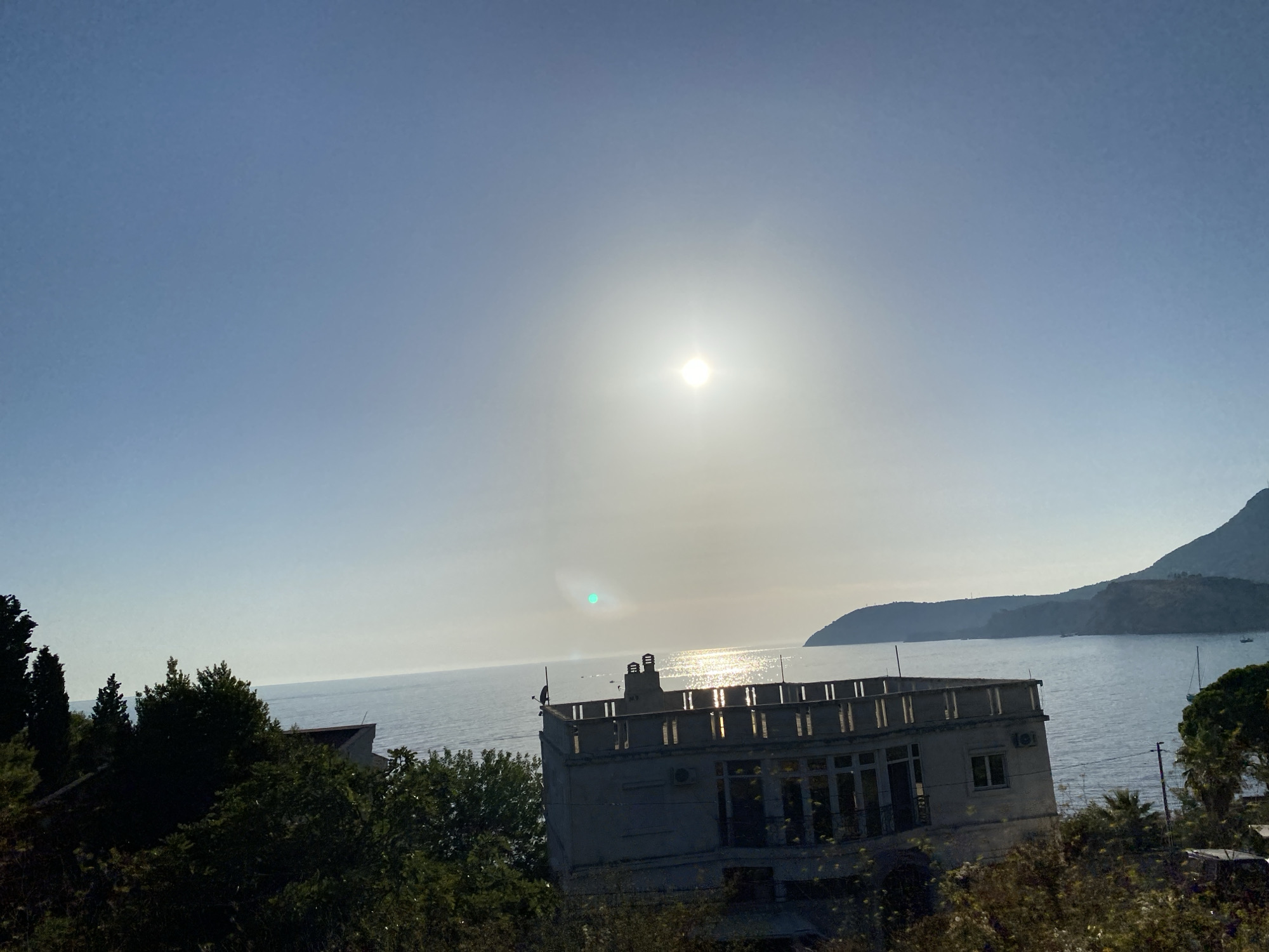 The image captures a view of the Adriatic Sea near Sutomore, Montenegro. The sun, high in the sky, casts a bright glare on the water, creating a shimmering effect. In the foreground, a building with a flat roof and a balcony overlooks the sea. Trees and foliage obscure parts of the building and the surrounding landscape. In the distance, hills and mountains form a backdrop to the scene. The sky is a clear blue, with some haze around the sun. A few boats are visible on the water.