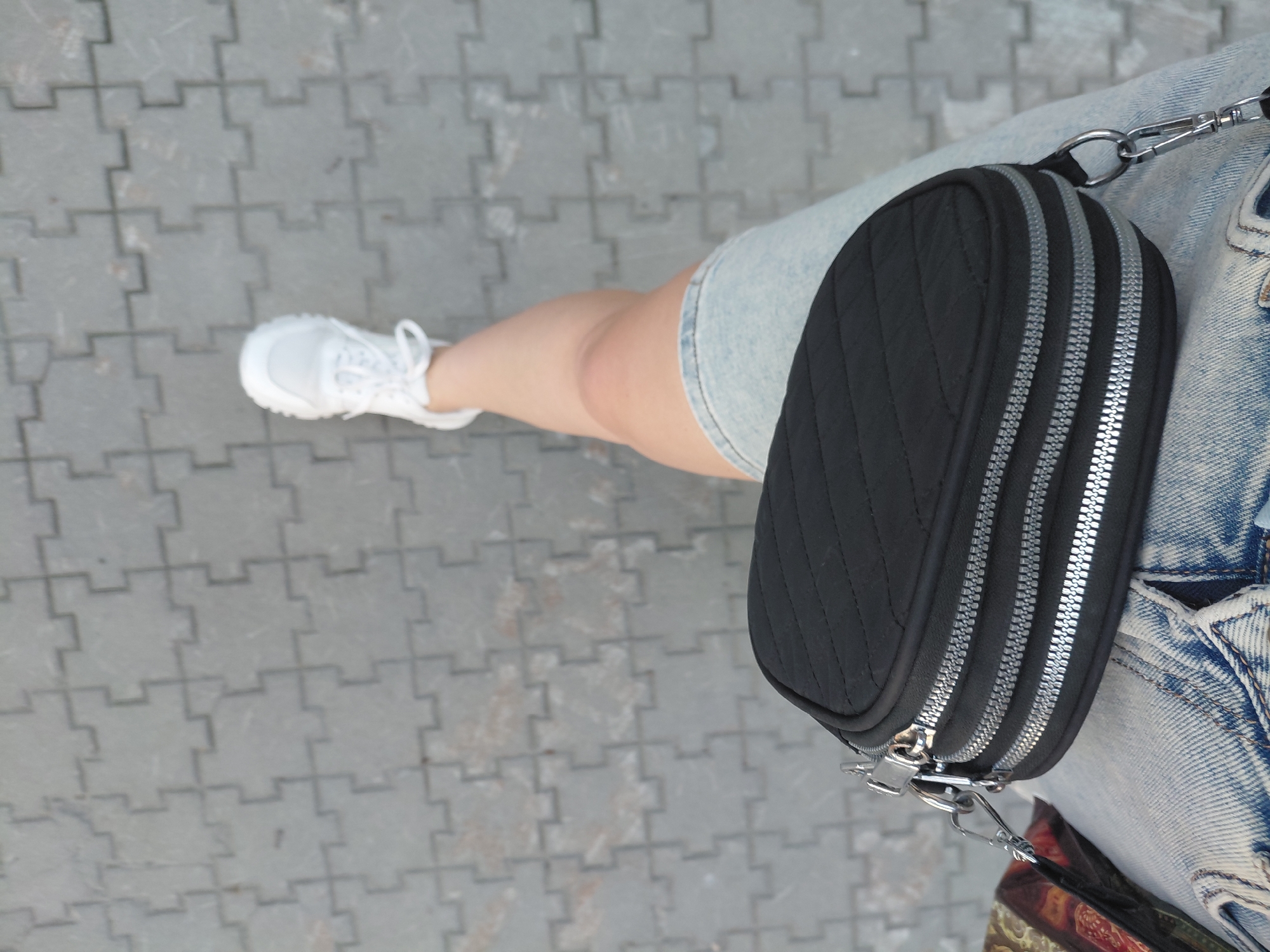 A person wearing white sneakers and a denim skirt is walking on a paved path. The path is made of grey interlocking stones. The person is carrying a black quilted crossbody bag with three zippers. A portion of another bag with a colorful pattern is visible on the left side of the frame. The image is taken from a high angle, looking down at the person's legs and the ground. No specific landmarks or identifying features of Novosibirsk, Russia are present in the image.