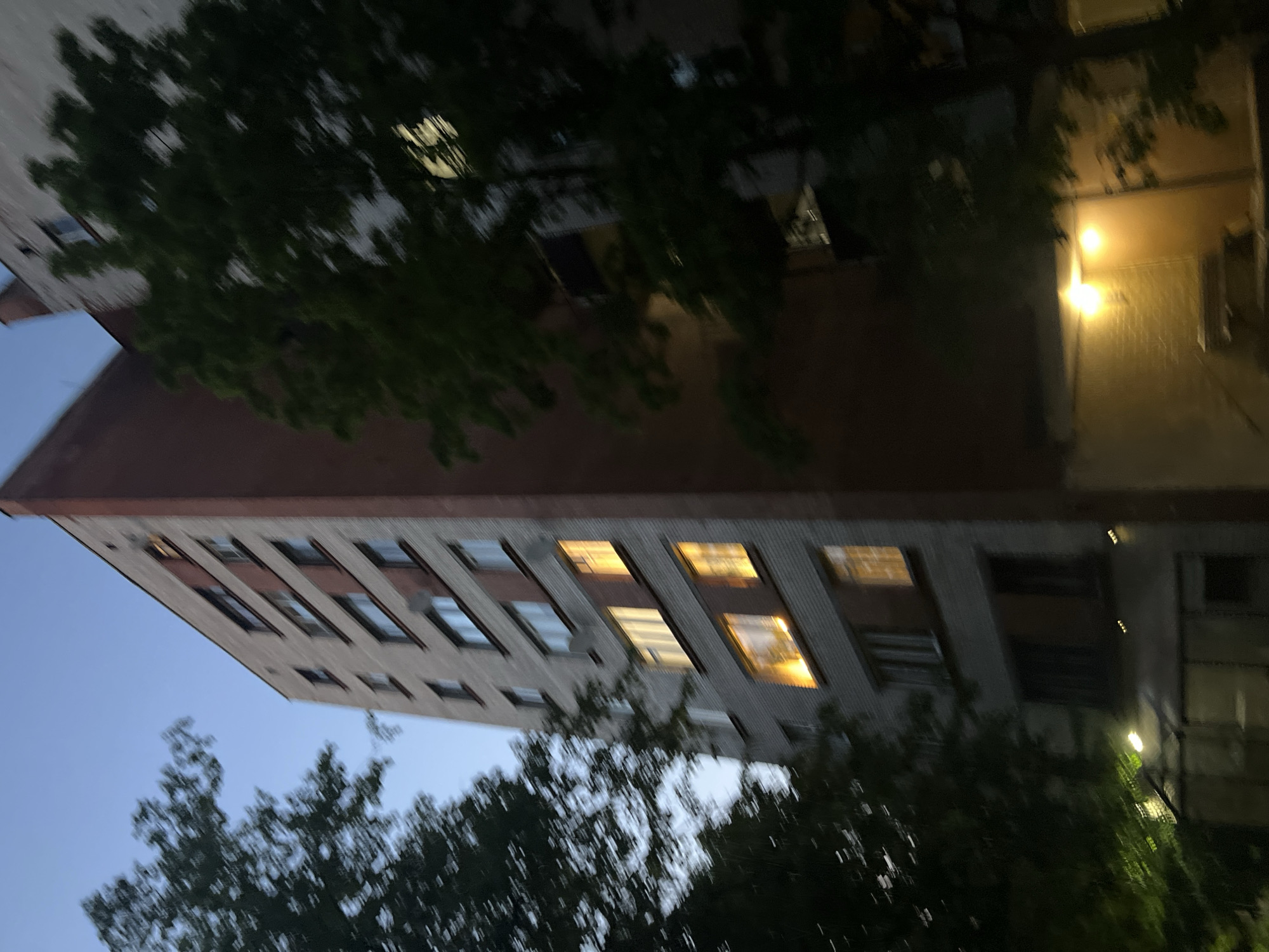 A multi-story residential building, approximately ten stories in height, is depicted at dusk or early night in Unknown, Russia. The building features a facade of light grey brickwork interspersed with reddish-brown sections, particularly around windows and on the right-side wall. Numerous rectangular windows are present, with several on the middle and lower floors emitting warm yellow interior light. The top left sky is a clear twilight blue. Dark green foliage from several trees partially obscures the building on both the left and right sides, exhibiting motion blur. At ground level on the right, two warm yellow exterior light fixtures illuminate a section of a yellowish wall and steps. The building's base also includes a large glass-fronted section, possibly an entrance or commercial space. No people or discernible actions are visible.
