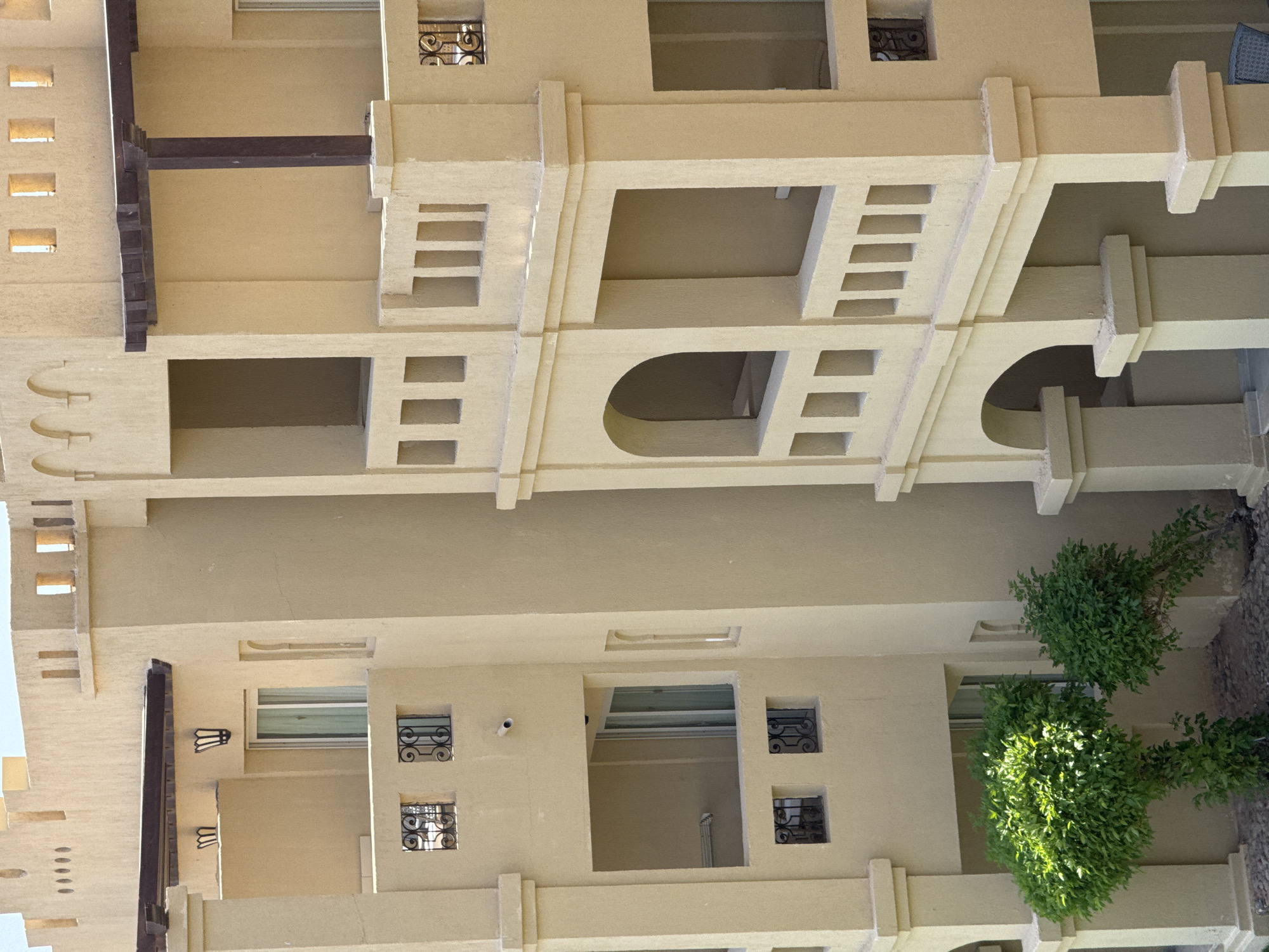 The image shows a side view of a beige apartment building in an unknown city in Egypt. The building has multiple levels with balconies and windows. Decorative elements include arched doorways and small, square windows with wrought-iron grates. There are also wooden beams supporting some of the balconies. Two small, green bushes are visible at the base of the building. The sky is visible in the upper portion of the image, appearing clear and blue. The architecture suggests a warm climate, possibly a resort or residential area.