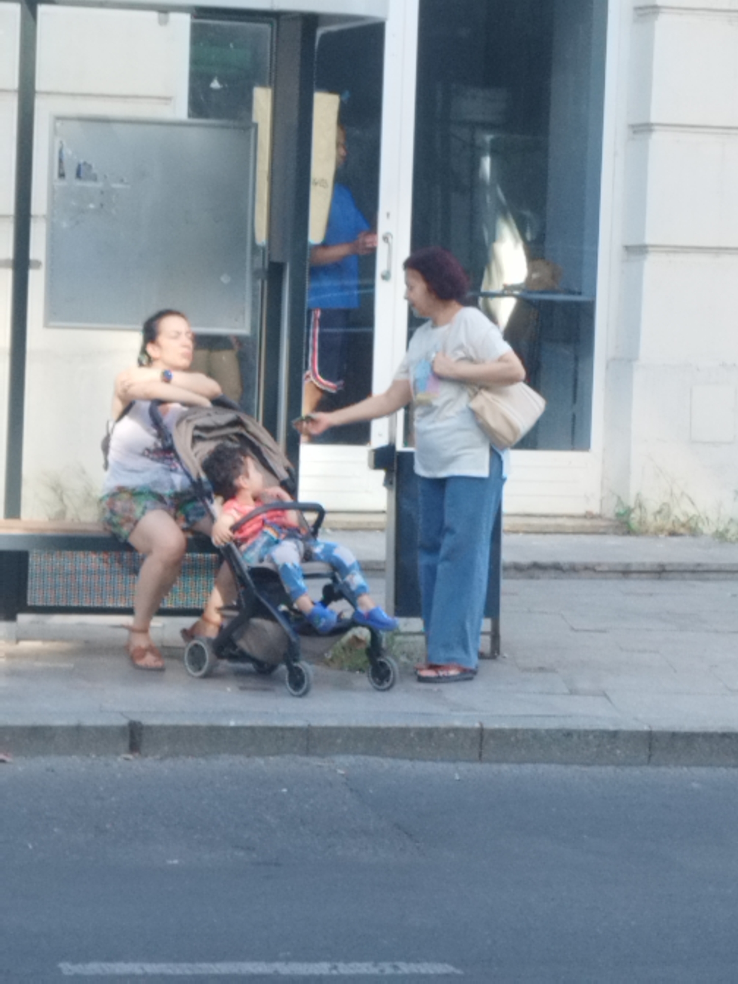 In Bucharest, Romania, on a sunlit sidewalk adjacent to a building with a glass-fronted entrance, a woman sits on a bench, holding a baby in a stroller. She wears a patterned skirt and a sleeveless top. To her right, another woman in blue pants and a white t-shirt stands, holding a beige purse over her shoulder. The baby in the stroller is wearing a red and blue outfit. The scene suggests a moment of pause or waiting on a public walkway.