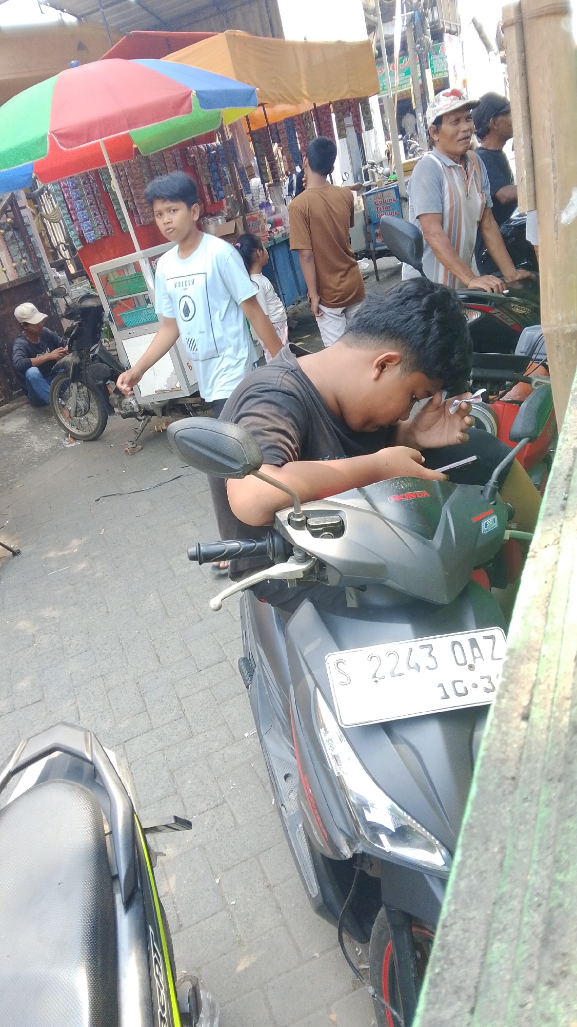 A boy in a black t-shirt is leaning over a black and gray motorcycle, looking down at his hands. He is holding a white object, possibly a cigarette or vape, to his mouth with his right hand and appears to be interacting with it. The motorcycle has a license plate that reads "S 2243 OAZ 10-23". In the background, there are several market stalls with colorful umbrellas. A young boy in a light blue t-shirt with a "Volcom" logo is walking away from the camera. Another person in a brown t-shirt is standing behind him. Further back, a man in a striped shirt and a cap is looking to the left. A black motorcycle is parked to the left of the scene. The ground is paved with gray bricks. The scene appears to be outdoors in a bustling market environment. Based on the license plate format and common market scenes, this location is likely in Mancar, Indonesia.