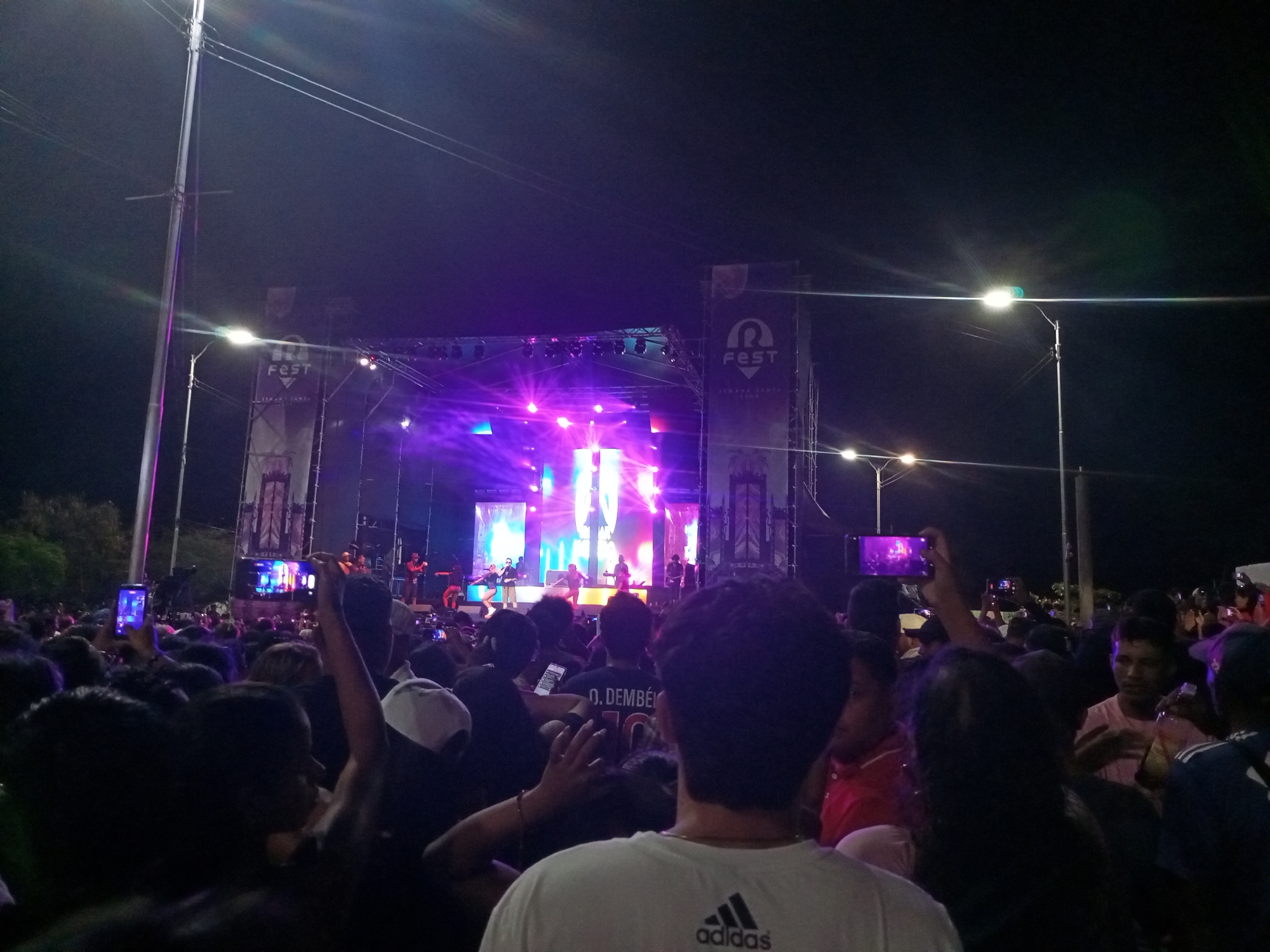 The image depicts a live concert taking place at night, likely at an outdoor venue in Porlamar, Venezuela. A large crowd of people is gathered, facing a brightly lit stage where a band is performing. Stage banners with "FEST" are visible. Many attendees are holding up their smartphones, recording or taking pictures of the event. The environment is dark, illuminated by stage lighting and streetlights. The crowd is dense, with people closely packed together. Some individuals are wearing casual clothing, with an Adidas t-shirt visible. There's evidence of tree cover on the left side of the frame.