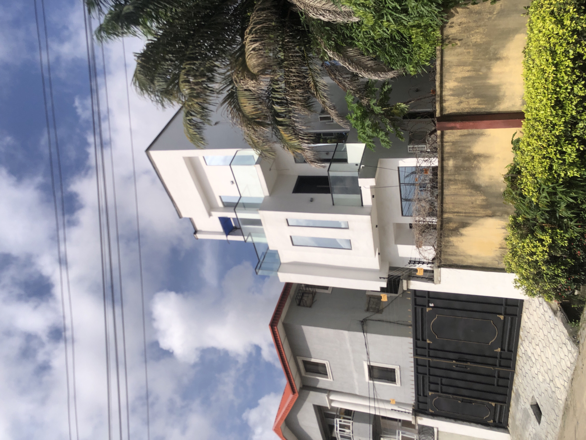 This image, taken in Ado, Nigeria, depicts a residential street scene on a bright, sunny day with a blue sky and scattered white clouds.
The primary subject is a modern, multi-story white building with a contemporary design. It features large, rectangular windows, some tinted, and glass railings on its balconies, giving it a sleek appearance. Lush green palm fronds partially obscure the upper part of this building and extend into the sky.
To the right of the white building, a yellowish-beige wall, partially covered by green bushes and foliage, separates properties. This wall shows some weathering and a darker, possibly reddish, horizontal beam embedded within it.
Adjacent to and partially in front of the white building, a second structure, appearing grey with a sloped orange-red roof, is visible. In front of this grey building, a tall, black metal gate with intricate patterns and gold-colored decorative accents stands prominently. A paved area, likely a driveway or sidewalk made of interlocking pavers, is visible in the foreground near the gate.
Overhead, utility lines stretch across the sky, suggesting the presence of power or communication infrastructure. The scene is static, capturing the architecture and immediate surroundings of the buildings. No people or overt activities are visible. The overall impression is one of a well-maintained, modern residential area in a tropical setting.
