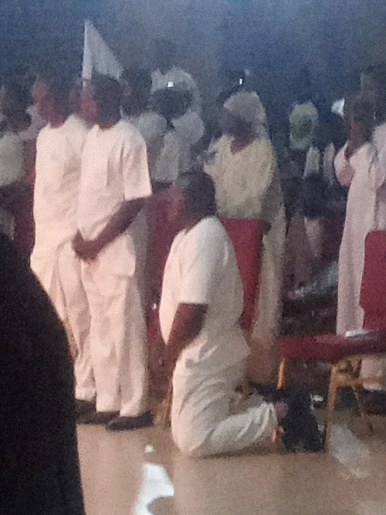 A group of people, appearing to be in prayer or contemplation, are gathered in what seems to be an indoor setting with muted lighting. The central figure is kneeling on the floor, facing away from the viewer, dressed in a white garment. Several other individuals in similar white attire stand behind the kneeling person, their faces partially obscured. The environment appears to be a public space, possibly a place of worship or a ceremonial gathering. There is a suggestion of seating or furniture, possibly red chairs, in the background. The image quality is low, with a grainy texture and soft focus, making precise identification of details difficult. The location is identified as Rukuba Road, Nigeria.