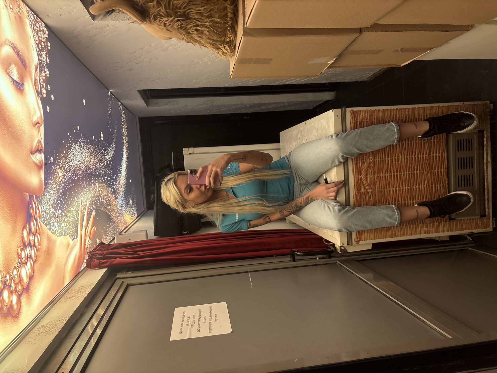 A woman with blonde hair is sitting on what appears to be an old refrigerator, taking a selfie with a pink phone. She's wearing a blue shirt and light-colored jeans, and has tattoos on her arms. Her legs are stretched out towards the right. The refrigerator is decorated with wicker and has an older-style metal ventilation grate. A poster with a woman's image and shiny details is on the wall to the left. A red drape is positioned below the poster. A sign with text in a foreign language is on the wall in the foreground. There are cardboard boxes stacked above the woman, partially obstructing the view of the ceiling. The location is unknown.
