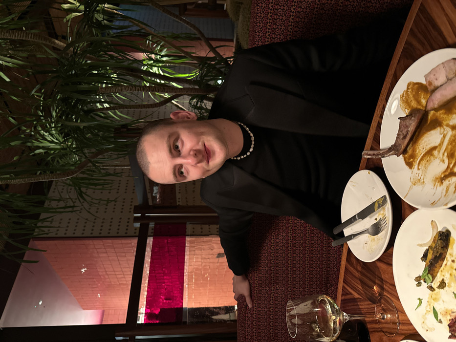 A man in a black blazer and pearl necklace sits at a table in a restaurant. He is smiling slightly at the camera. Next to him is a dark wooden table with several plates of unfinished food, including meat and sauce. Silverware is placed on one plate, and a partially filled wine glass rests on the table. A large potted plant stands to the left of the man, and a patterned wall and colored glass panels can be seen in the background.